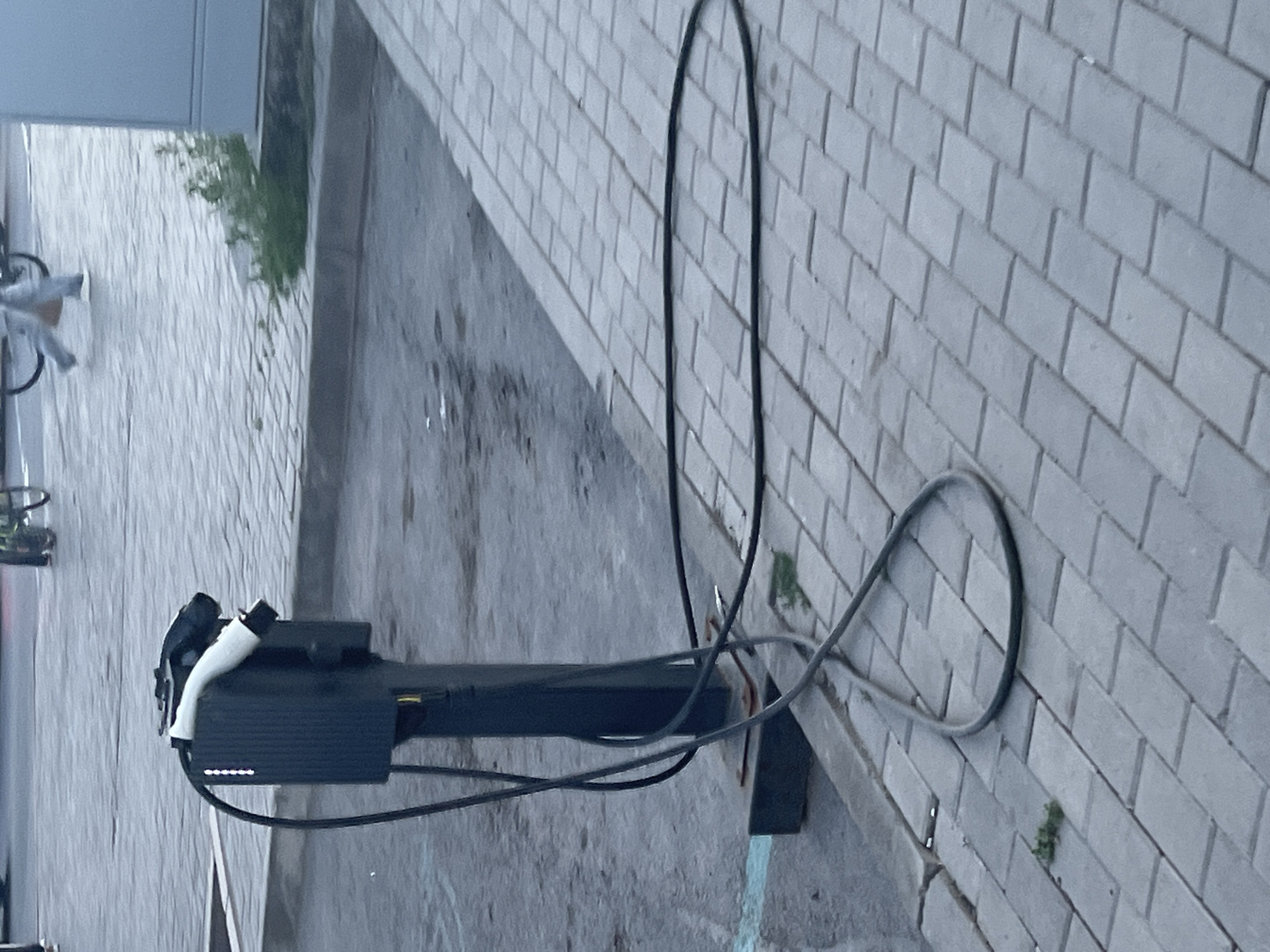 An electric vehicle charging station is situated on a paved walkway adjacent to a street, likely in Astana, Kazakhstan. The charging station, a black vertical unit with a white charging cable attached, is positioned near a curb. The street is wet, suggesting recent rain or street cleaning. In the background, a person walks past on the street, and a bicycle is visible. The overall scene is overcast, contributing to a muted color palette.