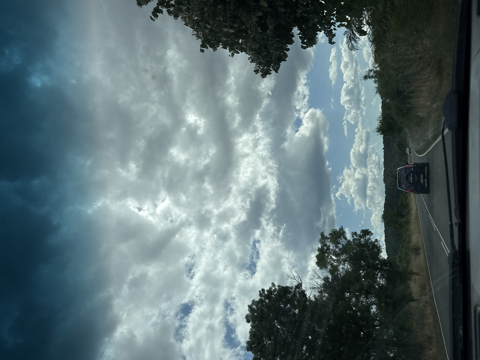 The image captures a scenic drive on a road, likely in or near Drachevo, Bulgaria, judging by the surrounding landscape. The perspective is from inside a vehicle, looking forward through the windshield. A dark-colored SUV, identified by "SUZUKI" on its spare tire cover, is the primary subject on the road, traveling in the same direction. Another vehicle is visible further ahead in the distance.
The environment is characterized by a winding asphalt road bordered by dense vegetation on both sides. To the left, large, dark green trees dominate the foreground, while the right side features a mix of tall grasses and smaller trees. Beyond the immediate roadside, rolling hills covered in lush green foliage form the background. The sky above is dramatic, filled with a mix of dark, moody clouds in the upper portion, transitioning to lighter, cumulus clouds scattered across patches of bright blue sky. The lighting suggests an overcast day with intermittent sunlight breaking through the clouds, creating a dynamic and atmospheric scene. The overall impression is one of a journey through a natural, somewhat wild, and picturesque landscape.