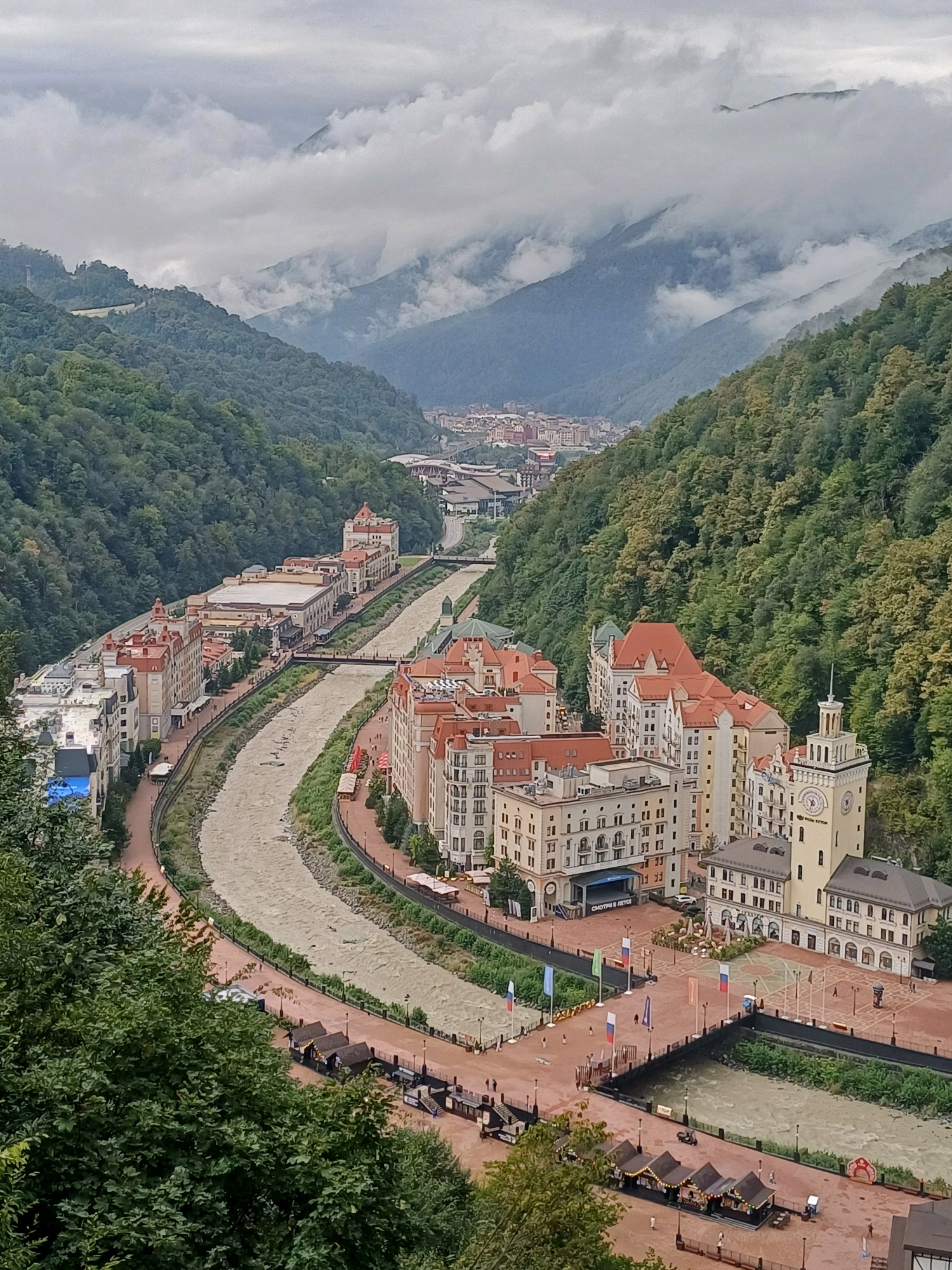 The image shows a wide, scenic view of a resort town nestled in a valley, with a river flowing through its center. The architecture is European-inspired, featuring multi-story buildings with red-tiled roofs and light-colored facades. Some buildings have clock towers and balconies, contributing to a picturesque and organized urban landscape. The river, which appears somewhat muddy, winds its way between the buildings, with bridges spanning it at various points.
The town is surrounded by lush, green mountains covered in dense forest. The sky is overcast with dramatic clouds, some of which are low-lying and partially obscure the mountain peaks, adding a sense of depth and atmosphere to the scene.
In the foreground and along the riverbanks, there are paved walkways and open public spaces. Several small, tent-like structures are set up, possibly for vendors or market stalls, with a few people visible walking around. Flags are also displayed in the public areas, suggesting a festive or organized event. The overall impression is of a well-developed tourist destination in a natural, mountainous setting. The presence of buildings, infrastructure, and recreational spaces indicates this is likely a resort or a town designed for tourism and leisure activities. The location is identified as Estosadok, Russia.