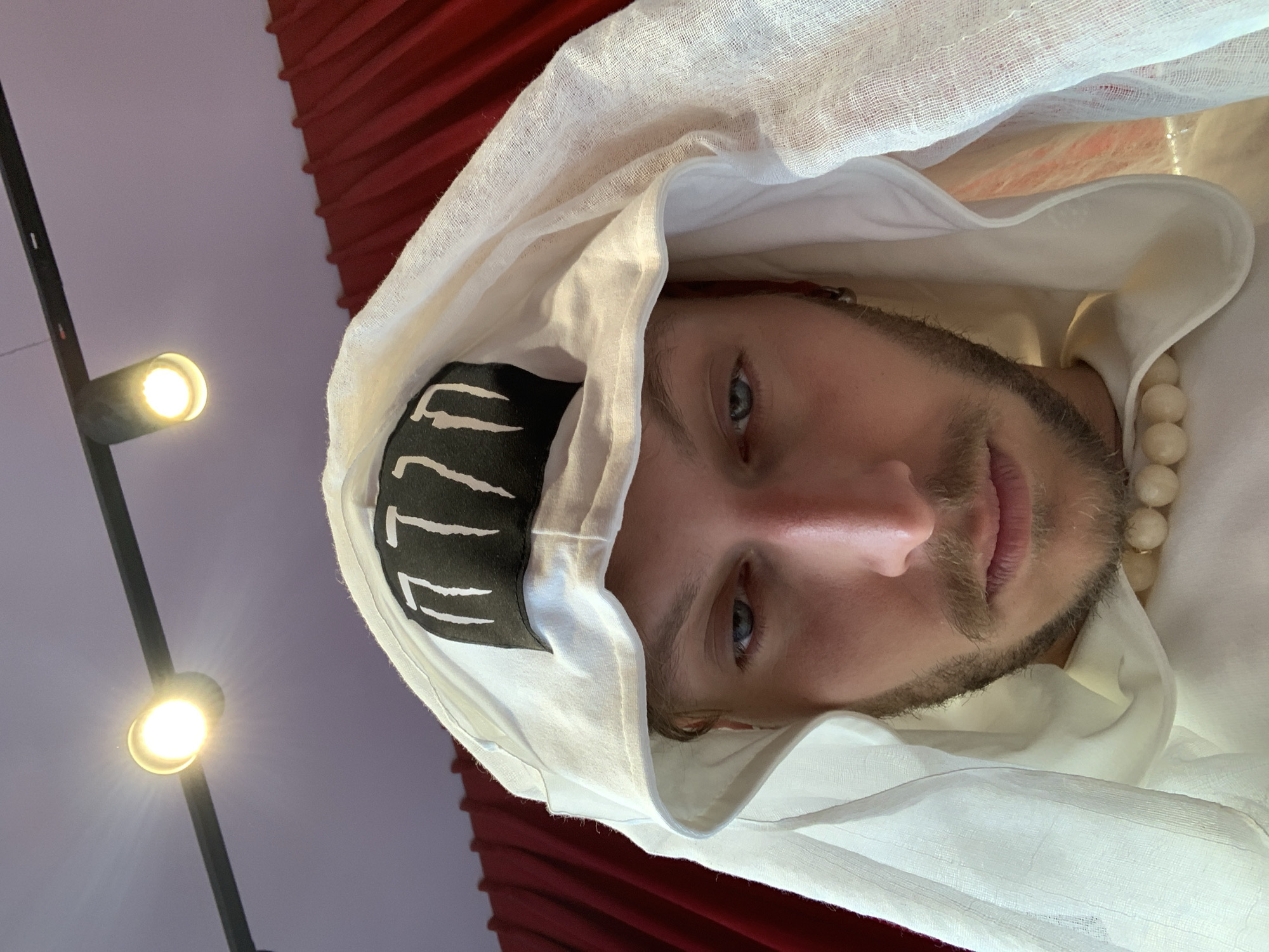 A close-up, rotated portrait of a man with fair skin, blue eyes, and a light beard and mustache is shown. He is wearing a white, draped head covering that obscures part of his forehead, with a black band visible underneath, decorated with white, vertical lines. A string of round, off-white beads is visible near his neck. The background includes a section of red drapery and a lighting fixture with two small, bright lights mounted on a black track. The location cannot be determined based solely on the image. The location of Tel-Aviv, Israel is extraneous and cannot be confirmed from the visual information.