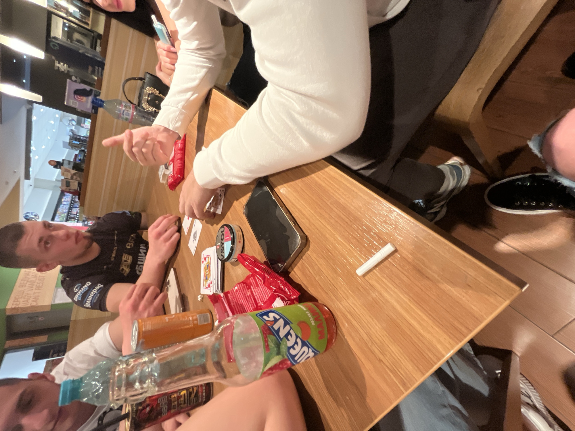 The image shows a group of people gathered around a wooden table. There are several items on the table, including a smartphone, playing cards, a pack of cigarettes, an orange juice can, a bottle labeled "Queen's," and a plastic bottle filled with liquid. One person is pointing with their finger. Another person is wearing a black t-shirt. The context is likely a casual gathering, possibly a game of cards. There are indications that the location might be Burgas, Bulgaria, as the t-shirt and juice can appear to be from local companies. The specific street address or location in Burgas is not identifiable from the image alone.
