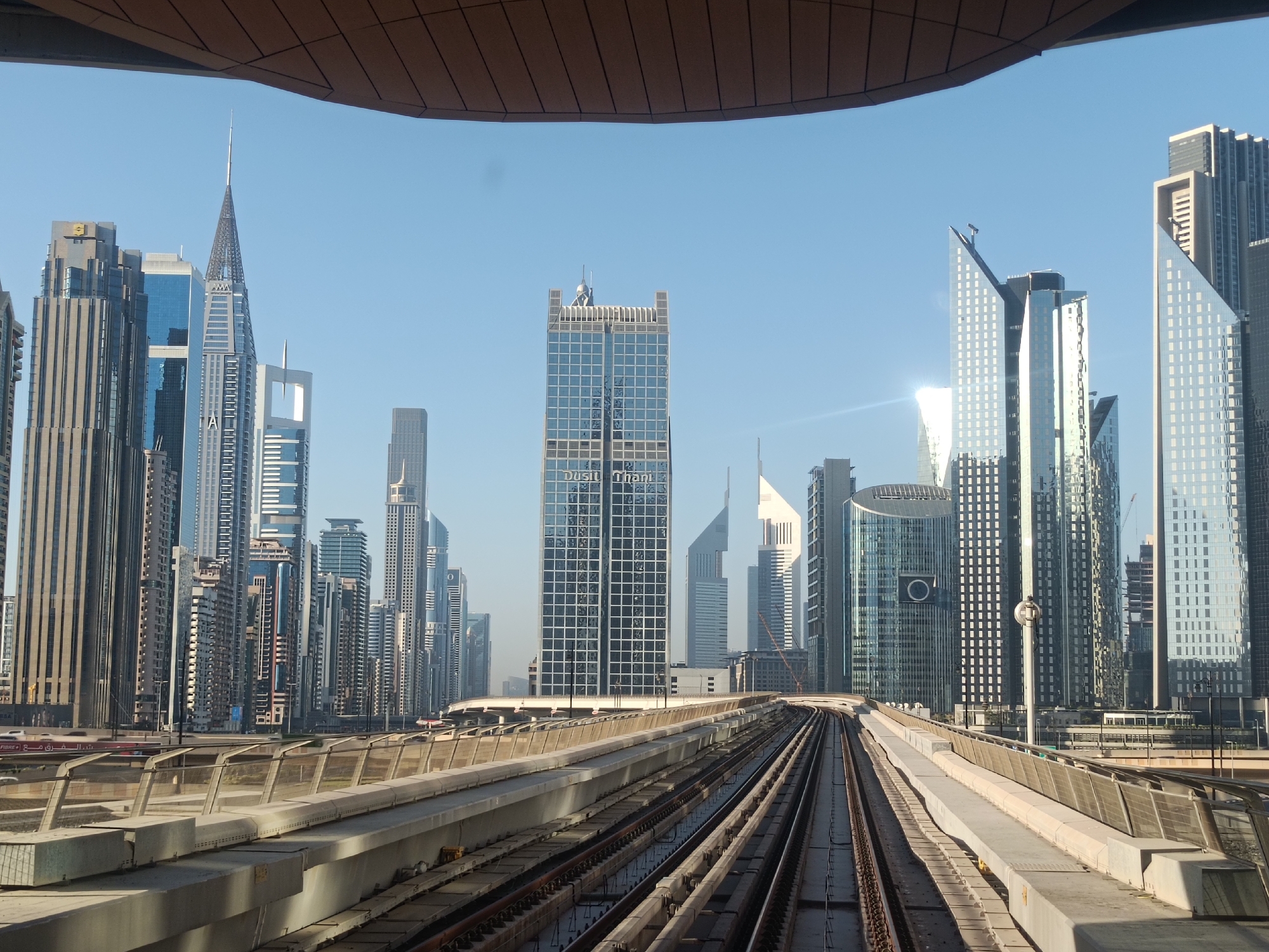 The image shows a view of the Dubai skyline in the United Arab Emirates from an elevated metro train. The foreground features the train tracks and supporting infrastructure. A series of modern skyscrapers dominates the cityscape. The building in the direct center of the frame features the words "Dusit Thani". The sky is clear and blue. The photograph is taken from inside the train, with a small portion of the roof visible at the top. No people are visible in the image.