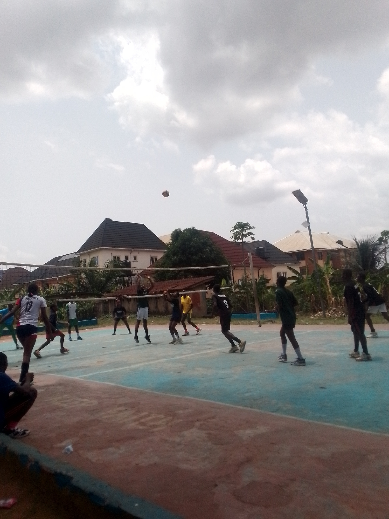 The image depicts a volleyball game in progress. The location appears to be an outdoor court with a blue and reddish-brown surface. A net divides the court. Several players are visible on both sides of the net, some are in the air attempting to hit the volleyball. The buildings and vegetation in the background suggest an urban area, with houses having multiple stories. A streetlight is also visible. The picture was taken in Onitsha, Nigeria.