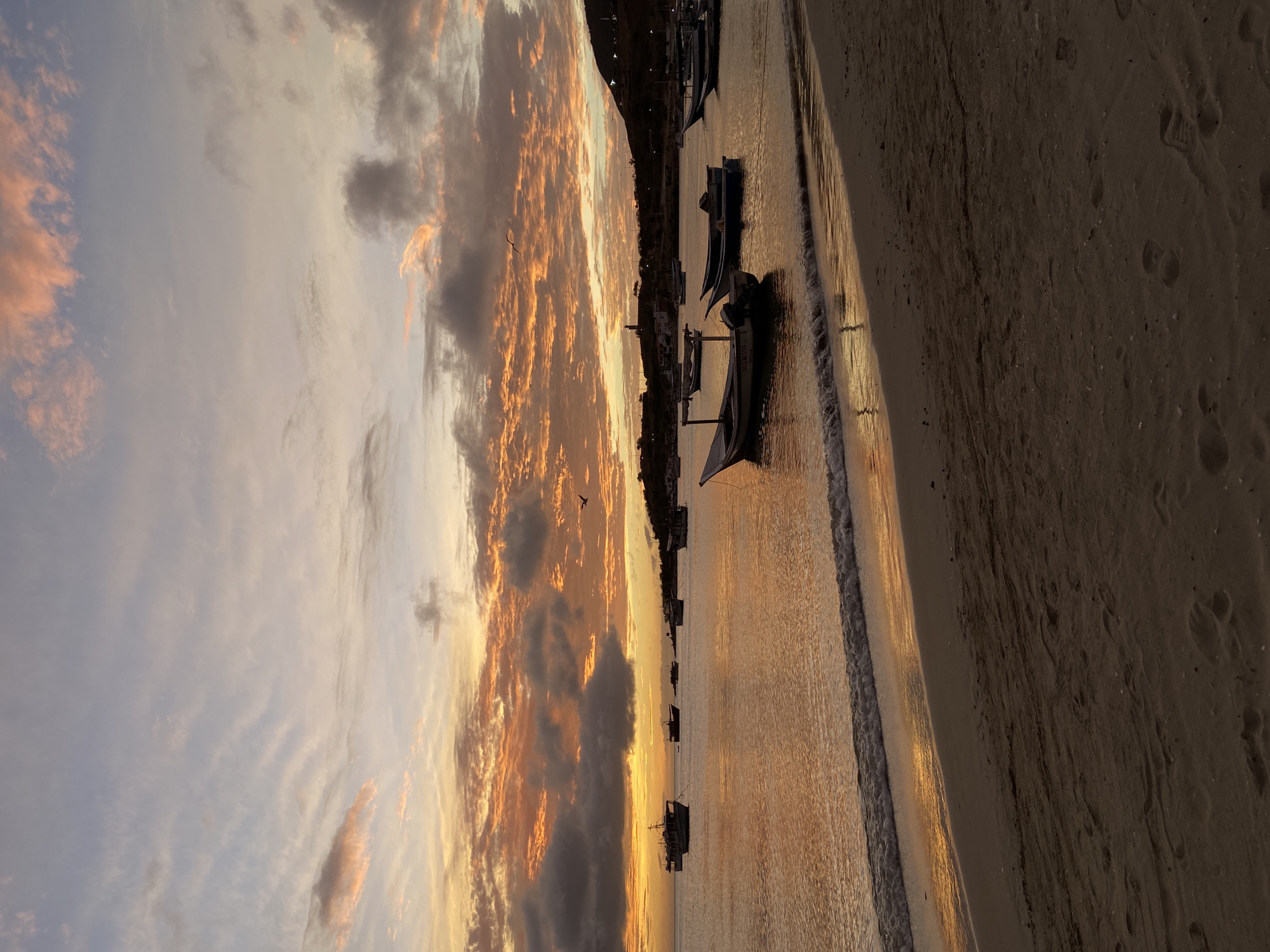 A wide shot captures a serene sunset over the waters of Juan Griego, Venezuela, with a sandy beach in the foreground. The sky is painted with vibrant hues of orange and pink clouds, interspersed with patches of pale blue and gray. The calm sea reflects the warm colors of the sunset, creating a shimmering effect. Several small boats are anchored in the water, their silhouettes dark against the bright sky. A gentle wave laps at the shore, leaving a wet line on the sand, which is imprinted with footprints. In the distance, a hilly coastline with some buildings is visible, bathed in the soft light of dusk. A lone bird is seen in flight against the dramatic sky.