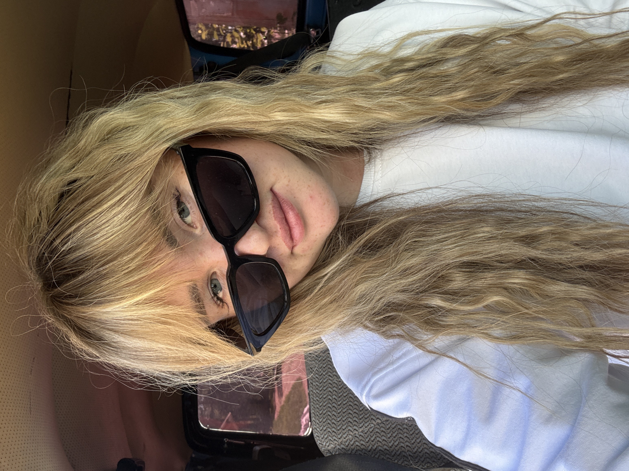 A woman with long, wavy blonde hair is wearing sunglasses and a white shirt. She appears to be inside a vehicle, possibly a car. The interior is a light brown or tan color. The background shows some out-of-focus objects, including what appears to be a window and perhaps parts of the vehicle's interior. The location is not discernible from the image.