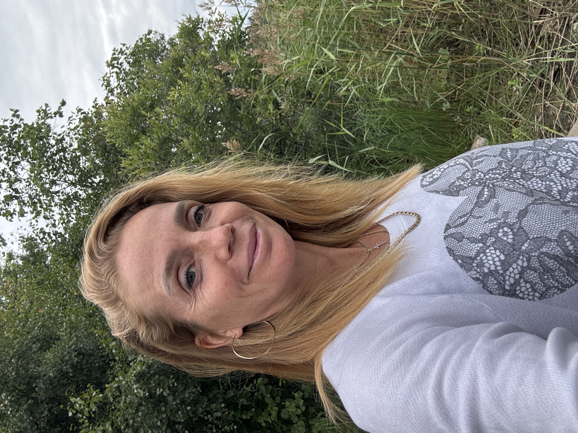 A blonde woman, appearing to be in her 40s, is captured in a close-up, outdoor selfie. She has fair skin with freckles, blue eyes, and light brown hair that falls past her shoulders. She is wearing a white crew neck sweater adorned with a large gray lace-like pattern on the front. She also sports a gold hoop earring in her left ear and a gold chain necklace with a small cross pendant. The woman is smiling, looking directly at the camera.
The background consists of lush green foliage and tall, dry grass, suggesting a natural, possibly marshy or riverside environment. The sky above is overcast with a light gray hue. The image was taken in Kulikovo, Russia.