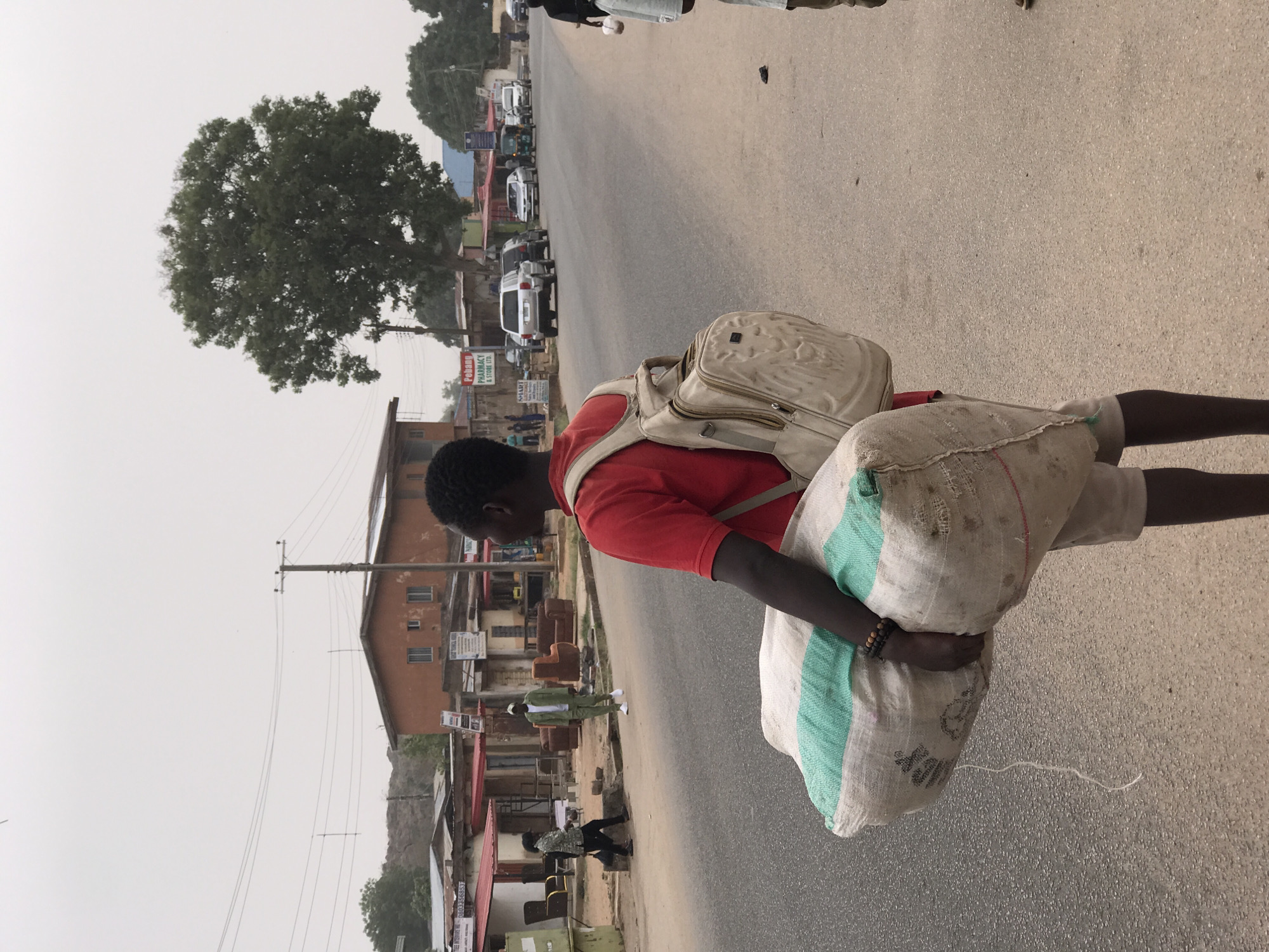 A man in Jos, Nigeria walks along a paved road carrying two large sacks. He is wearing a red shirt, light-colored shorts, and a backpack. The sacks appear to be heavy and worn. He wears a wristband on one arm. The road is lined with buildings and trees. Several cars are parked along the side of the road. Overhead power lines are visible. The sky is overcast and hazy.