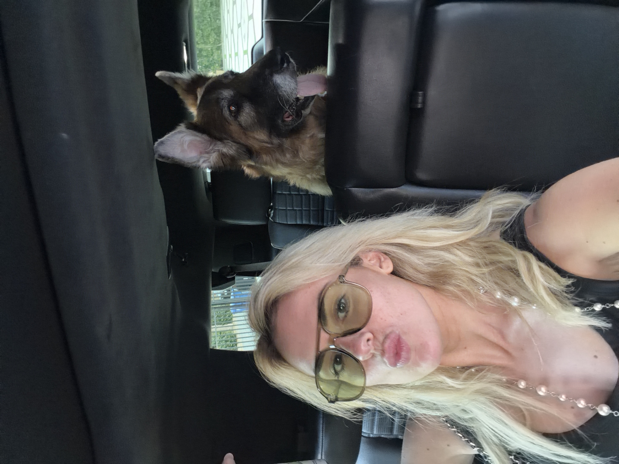 A woman and a German Shepherd dog are in the back of a black vehicle. The woman has long blonde hair and is wearing sunglasses and a pearl necklace. She is facing the camera with her lips pursed. The dog is behind the seat, panting with its tongue out. The location appears to be in a city, possibly Los Angeles, as indicated in the prompt. There are buildings visible through the back windows of the vehicle.