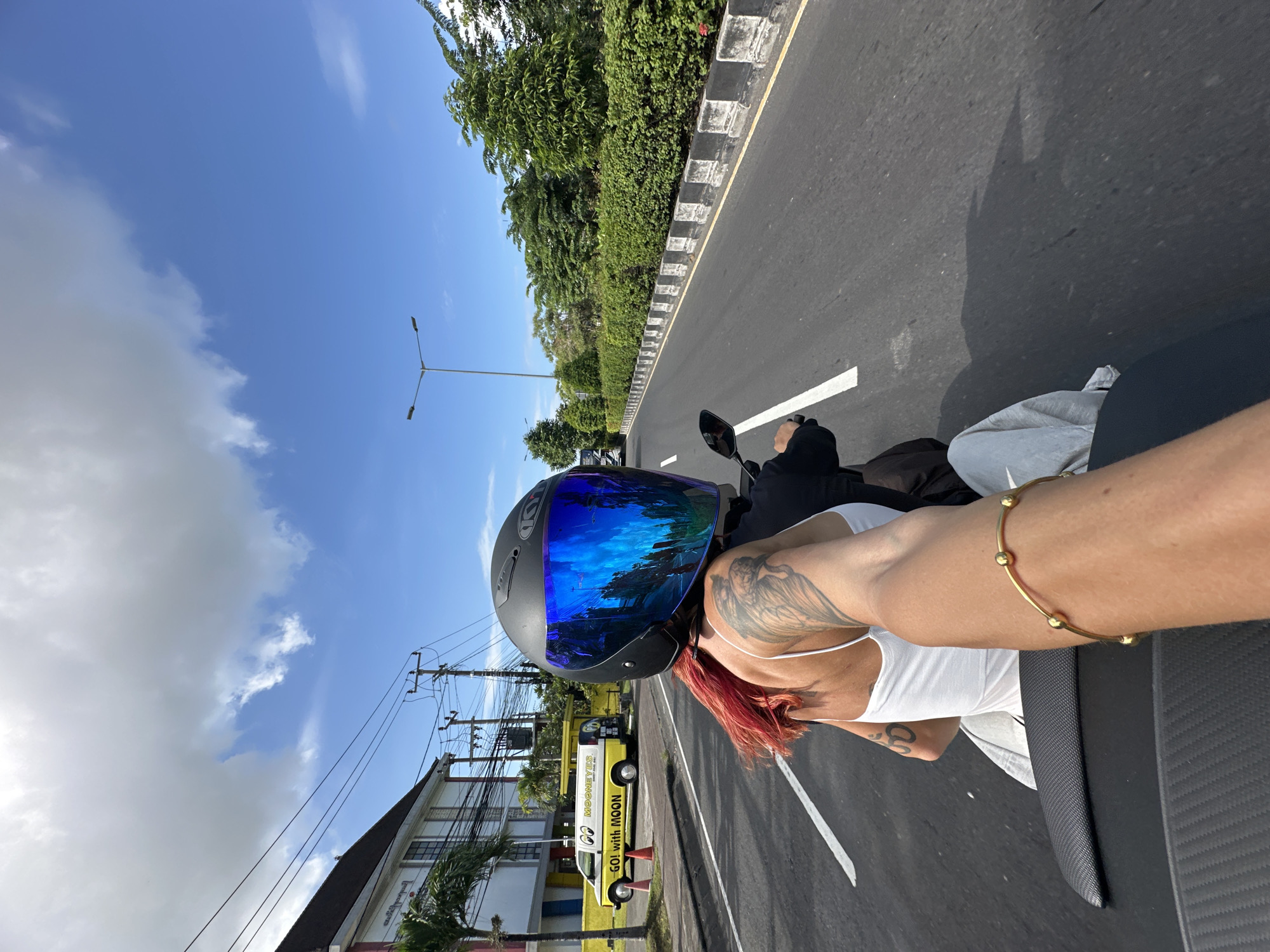 A person with red hair wearing a blue-tinted helmet, a white tank top, and a gold arm bracelet is riding a motorcycle on a paved road. The person has tattoos on their arm and shoulder. The road is divided by a solid white line and has a black and white checkered border. Lush green trees line the roadside. In the background, there is a building and a yellow van with the words "MOONEYES" and "GO! WITH MOON" written on it. The sky is bright blue with scattered white clouds. The image likely originates from the city of Kuta, Indonesia.