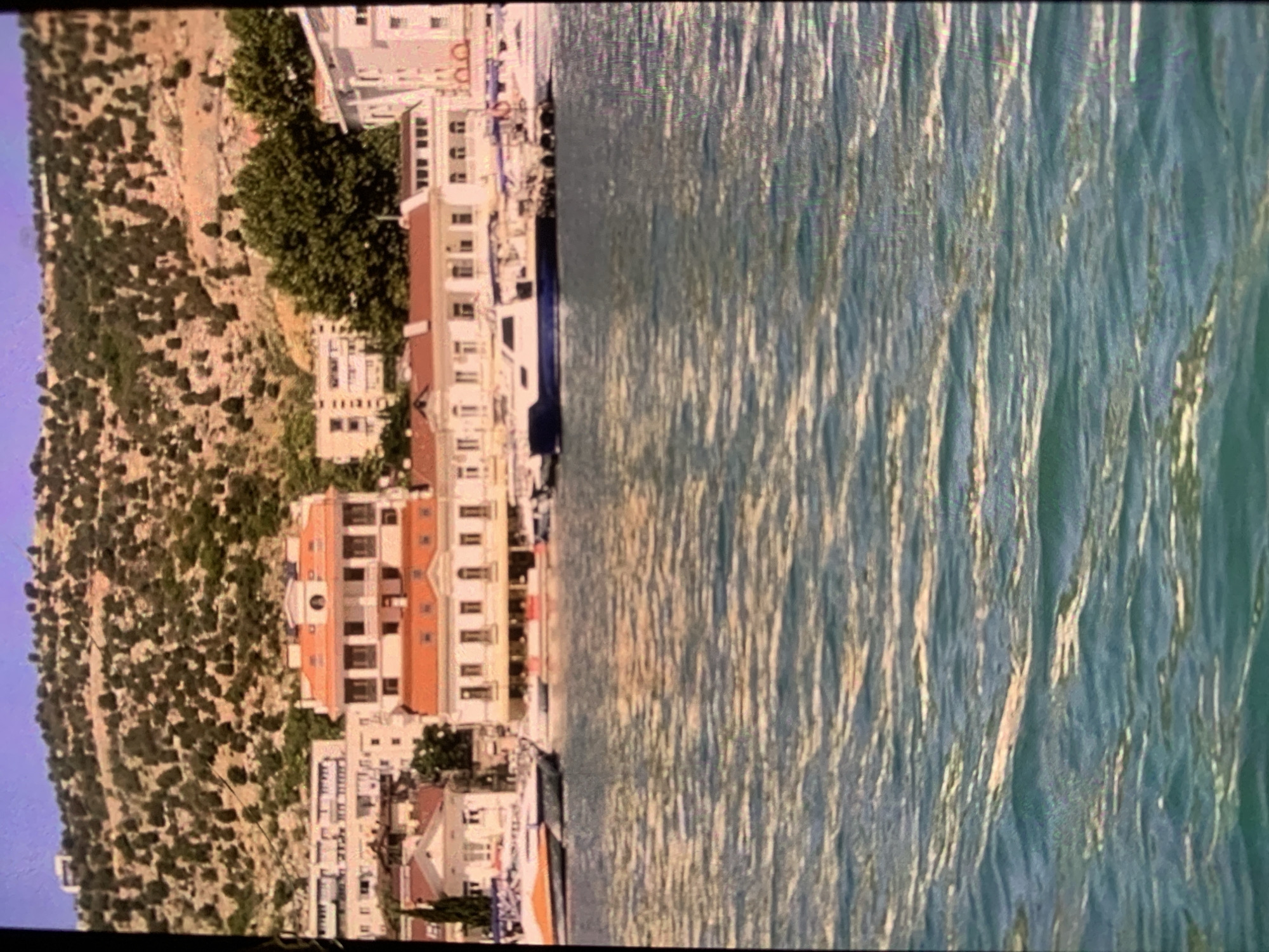 The image shows a waterfront in Sevastopol, Ukraine, with several buildings lining the shore. In the foreground, the water is rippled and appears to be a greenish-blue color. Several boats are docked or cruising in the water. Behind the waterfront, a hillside is covered with sparse vegetation and trees, and more buildings are visible further up the slope. The sky is a clear, pale blue. The architecture of the buildings suggests a Mediterranean influence, with red-tiled roofs and light-colored facades. The scene depicts a typical coastal urban environment.