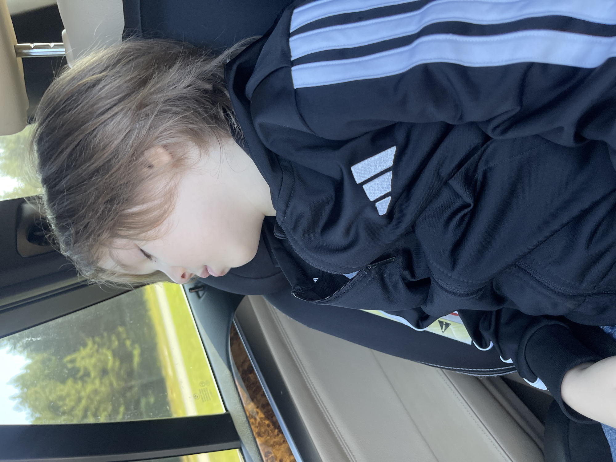 A young child with brown hair is sleeping in a car seat. The child is wearing a black Adidas tracksuit. The car interior is visible, with a light-colored seat and wood trim on the door. Outside the window, trees and a road can be seen. The scene is lit by natural sunlight. The image appears to have been taken during the daytime in a suburban or rural area.