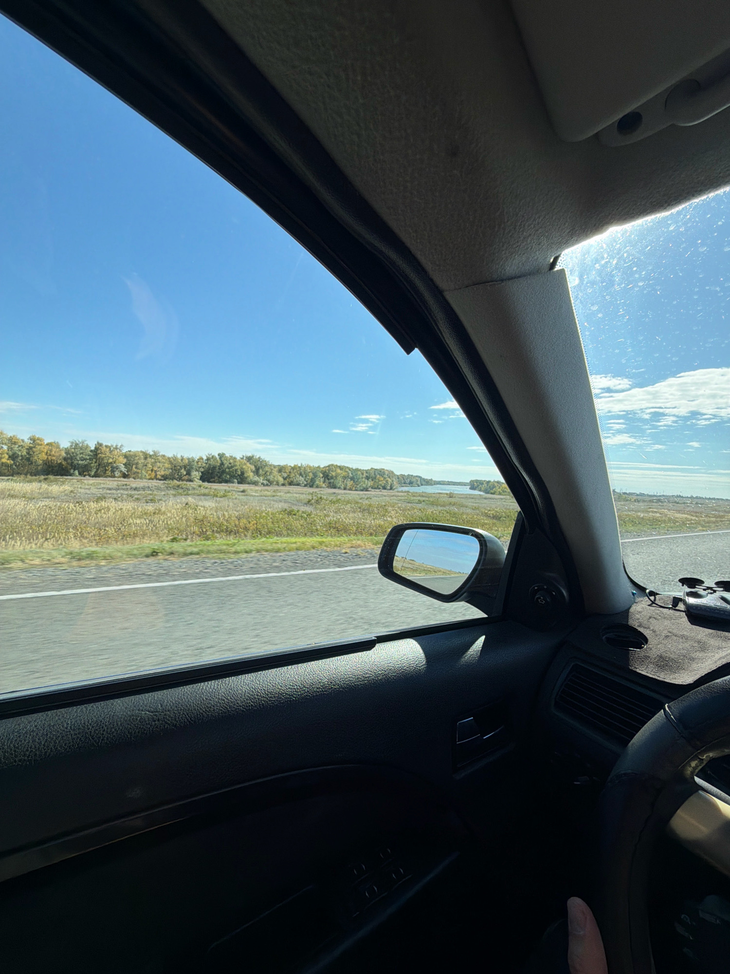 The image is taken from inside a car, looking out the passenger side window towards a landscape under a bright blue sky with scattered clouds. The car's interior is visible, including the door panel, window frame, and part of the dashboard and steering wheel. Outside the window, a road with a white line runs alongside a field of dry, golden grass and scattered green bushes, leading to a body of water, possibly a river or lake, bordered by a line of trees with autumn foliage. The scene is one of travel through a rural, possibly steppe-like environment. The location is identified as Көктүбек ауылдық округі, Kazakhstan.