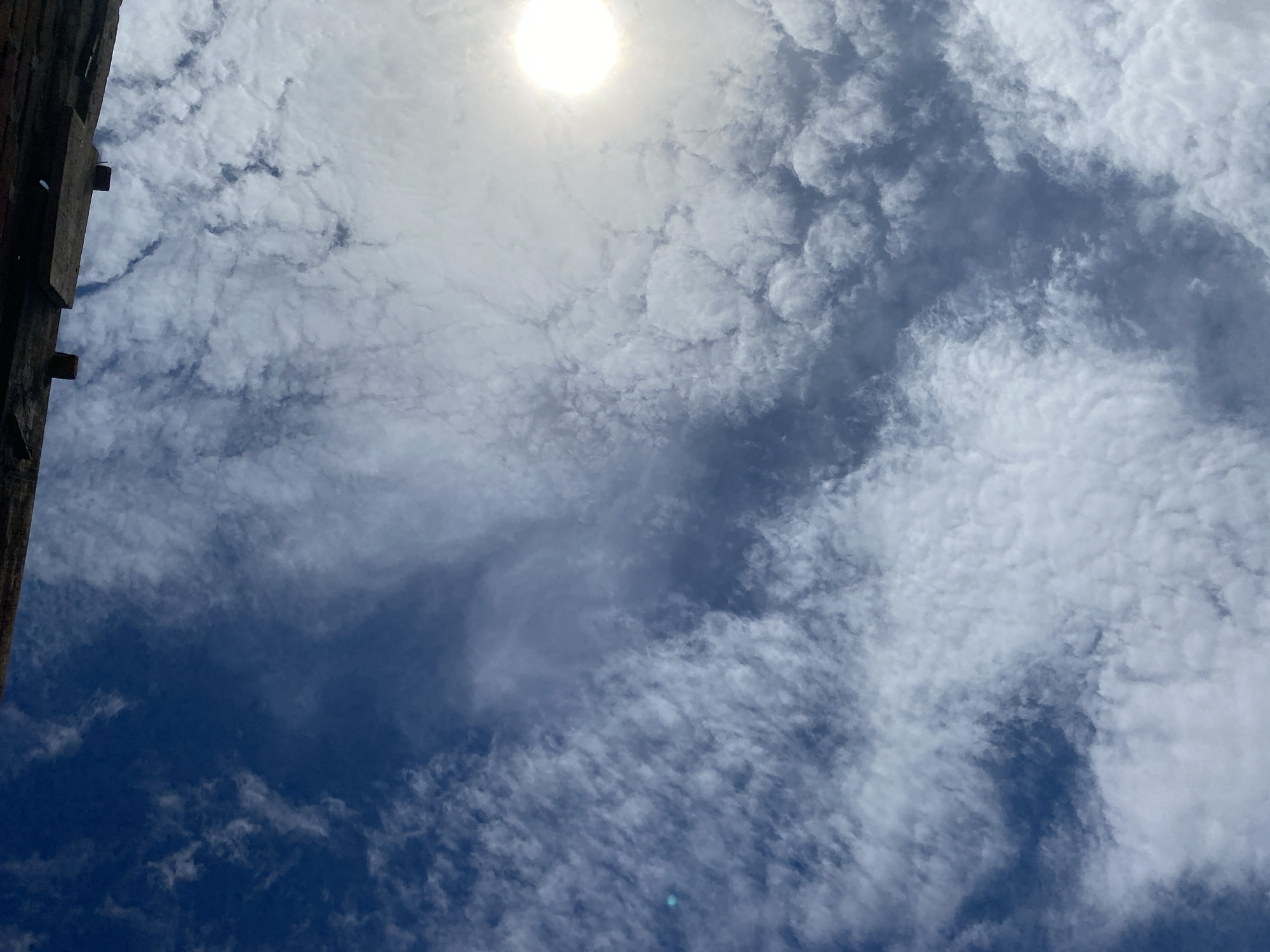 A low-angle, vertical shot captures a vibrant blue sky filled with an assortment of white, wispy clouds, and a bright sun shining on the upper right side. The sun, a brilliant orb, is partially obscured by the clouds, creating a hazy effect. In the upper portion of the image, a dark, angled structure, possibly a wooden beam or a section of a roof, juts into the frame, casting shadows. The clouds are varied in texture, with some appearing fluffy and dense, while others are thin and stretched across the sky. The overall impression is one of a bright, clear day with a dynamic cloudscape.