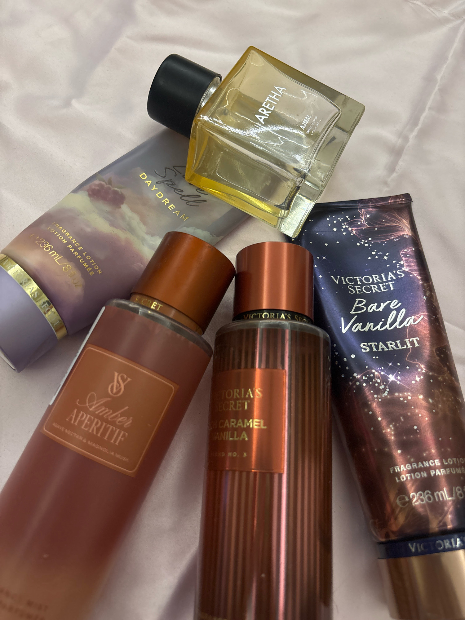 The image displays a flat lay arrangement of various fragrance products on a light pink, textured surface. The products include a clear glass perfume bottle with a black cap labeled "ARETHA" and "AJMAL," a purple tube of lotion with "DAYDREAM" and "VICTORIAS FOOT" visible, a bronzed spray bottle with "VICTORIAS SECRET Amber APERITIF" and notes of "AGAVE NECTAR & MAGNOLIA MUSK," another bronzed spray bottle with "VICTORIAS SECRET CARAMEL VANILLA," and a dark blue tube of lotion with "VICTORIA'S SECRET Bare Vanilla STARLIT" and a sparkly design. The surface appears to be fabric, possibly a bedspread or tablecloth. There are no people, actions, or interactions depicted in the image. The lighting suggests an indoor setting, and there are no visible landmarks or environmental cues to indicate a specific location in Jaipur, India.