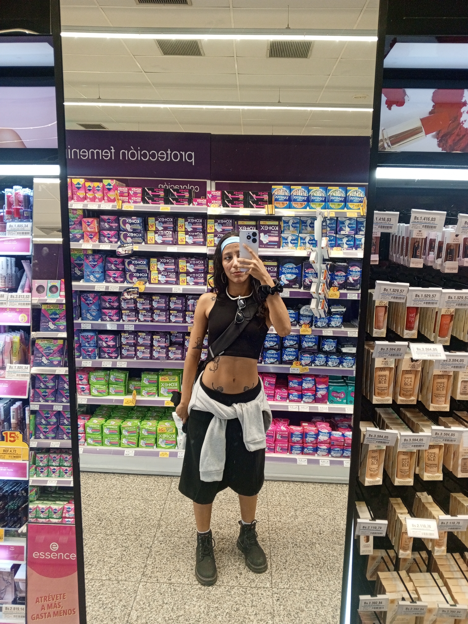 A woman stands in front of a mirror in what appears to be a supermarket aisle. The aisle is filled with feminine hygiene products and cosmetics. The woman is wearing a black crop top and black shorts, and a white sweater is tied around her waist. She is holding a smartphone in her left hand, possibly taking a selfie. The shelves behind her are stocked with products from brands like Kotex and Essence. Price tags in Bolivars (Bs) are visible on the shelves. Based on the prices shown, this might be a supermarket in a location where hyperinflation is present. The location of Pampatar, Venezuela is not directly ascertainable from the image.