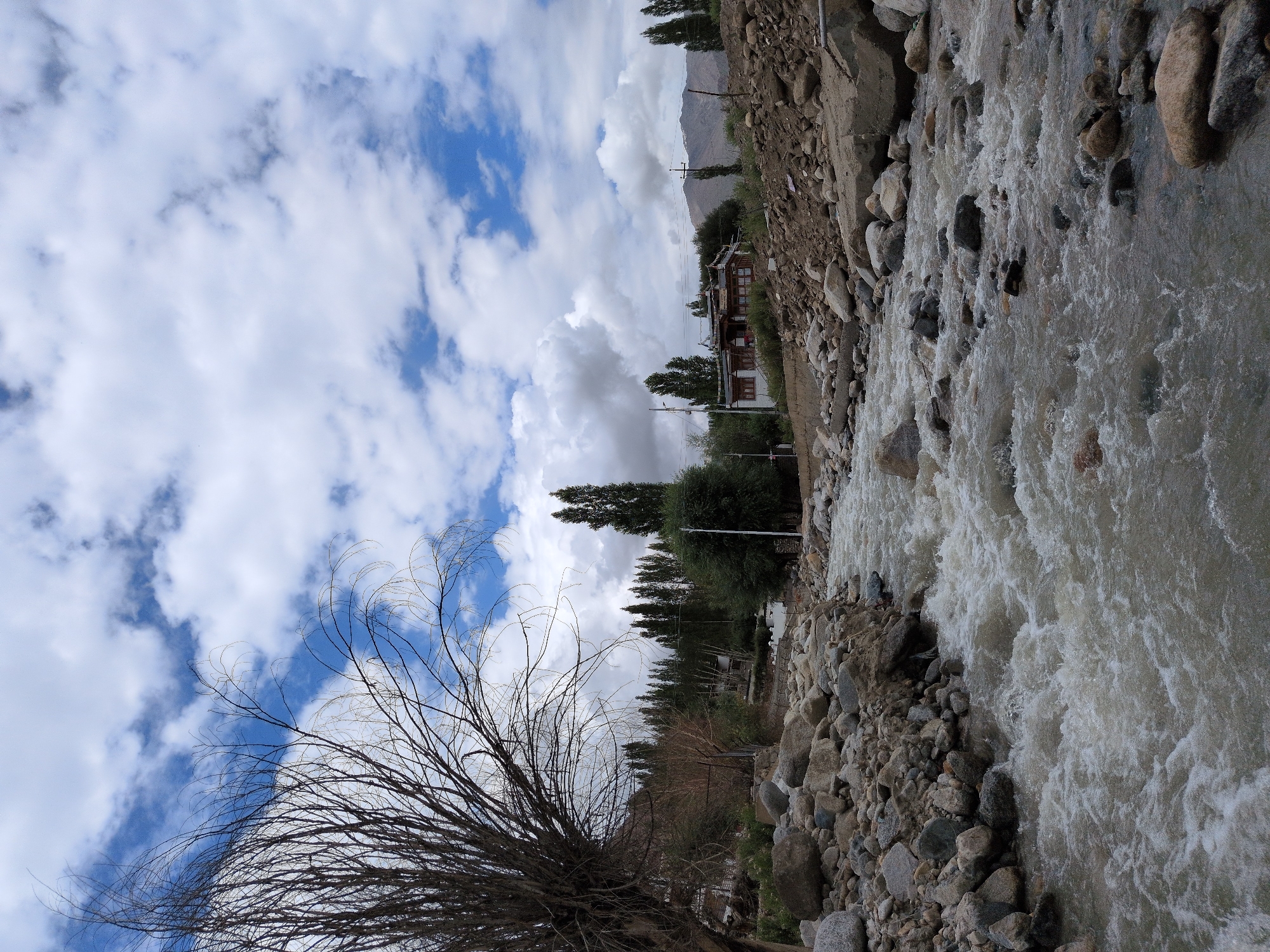 The image depicts a scenic view of a flowing river in Leh, India. The river, with its swirling currents and white foam, dominates the foreground, bordered by a rocky embankment on the left. In the mid-ground, a cluster of trees and a traditional Leh-style building with a brown roof are visible. The building appears to be situated on a slightly elevated area overlooking the river. The background features rolling hills and a vast sky filled with dramatic white clouds against a blue backdrop. The overall environment suggests a natural landscape with some human habitation.