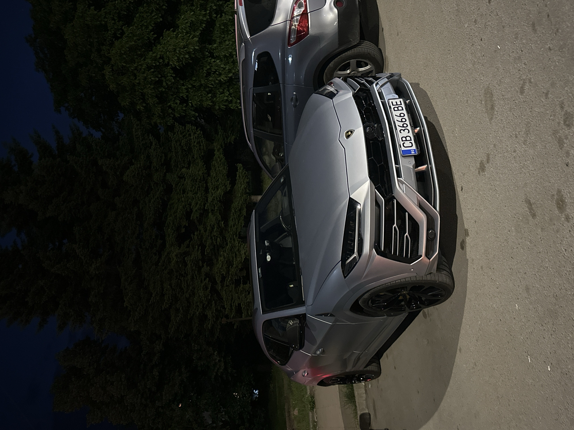 A gray Lamborghini Urus and a gray Nissan Qashqai are parked on a paved street. The Lamborghini has a Bulgarian license plate, "CB 3666 BE". A dark, heavily foliated area, possibly a park or wooded area, is adjacent to the street. The image was taken at night. The location is assumed to be Veliko Tarnovo, Bulgaria.