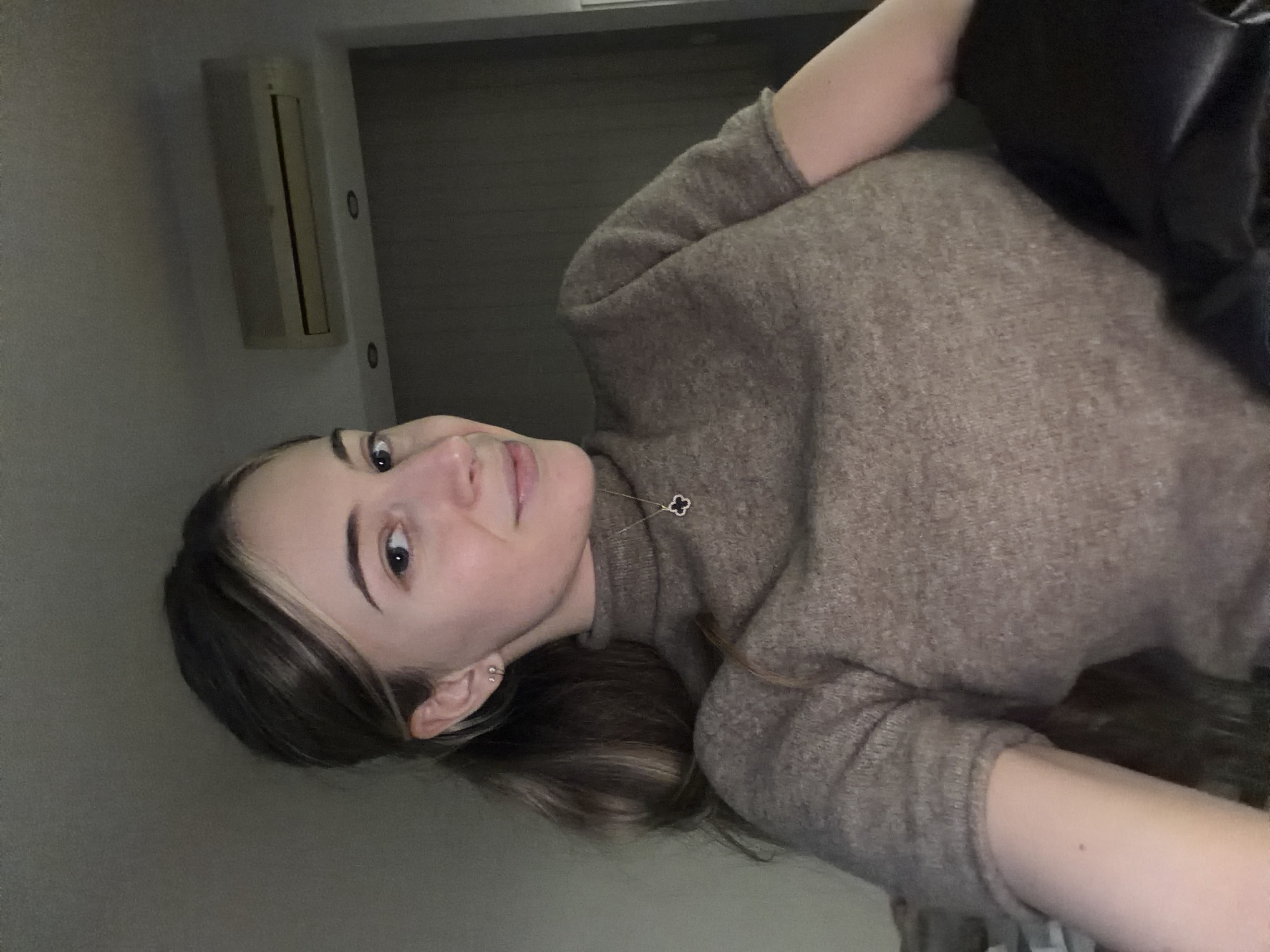 A young woman with light skin and dark, shoulder-length hair is seen in a close-up shot. She has an apparent lighter blonde highlight along her hairline. She is wearing a light brown knit turtleneck sweater. She has a clover-shaped pendant necklace. An air conditioning unit is affixed to the wall in the background. The exact location in the city of Plovdiv, Bulgaria cannot be determined from this image.
