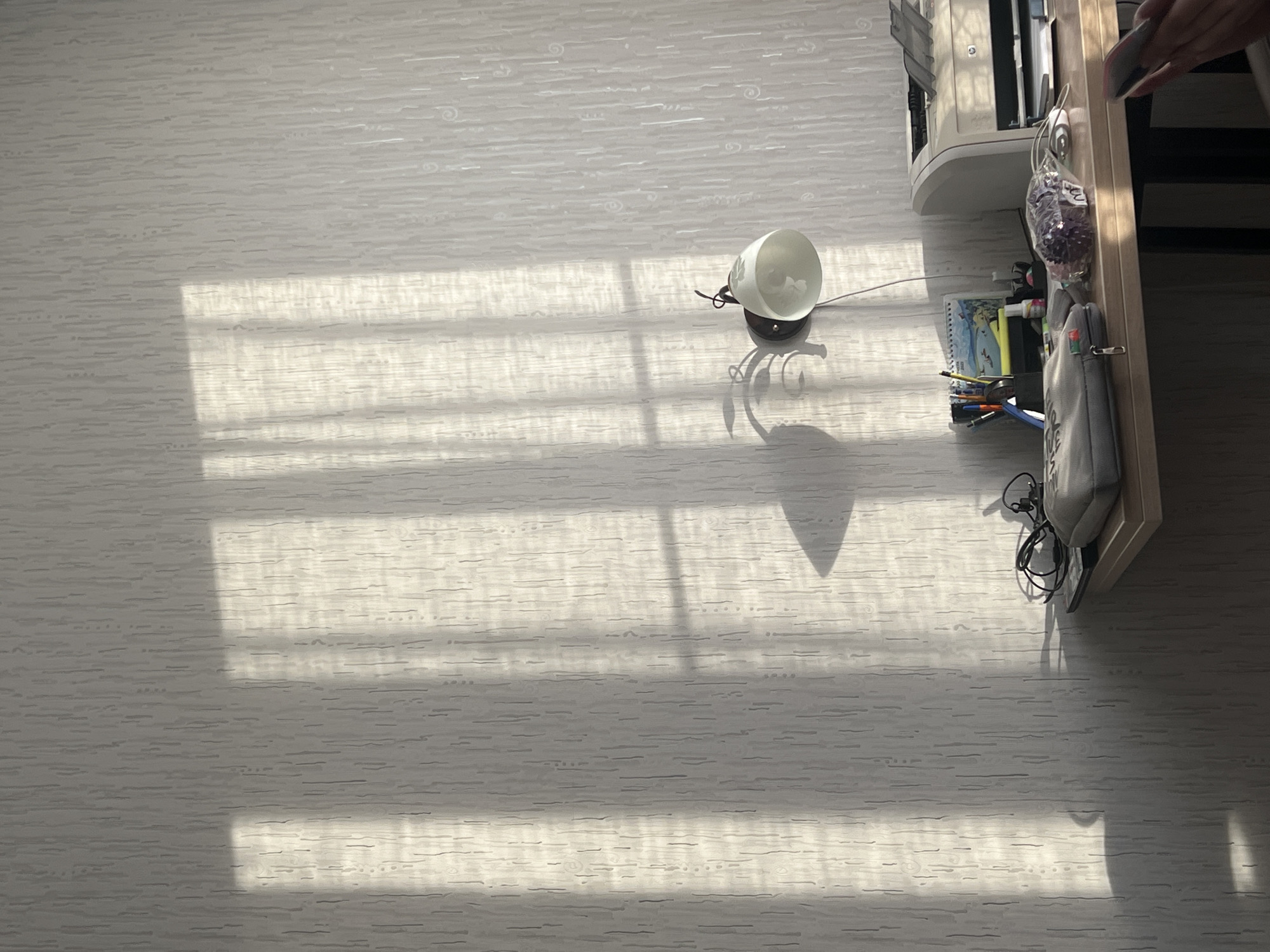 A high-angle shot captures a room, possibly in Perm, Russia, based on the user's prompt. The room appears to be illuminated by natural light casting distinct parallel shadows across a textured, likely wallpapered floor.
A lamp with a white, cone-shaped shade sits on the floor, its shadow also visible. Adjacent to the shadows and lamp, a shelf or desk edge is visible, upon which rests a printer, various writing implements (pens, pencils), notebooks, and a small bag. A hand holding a mobile phone or remote is visible in the right corner.