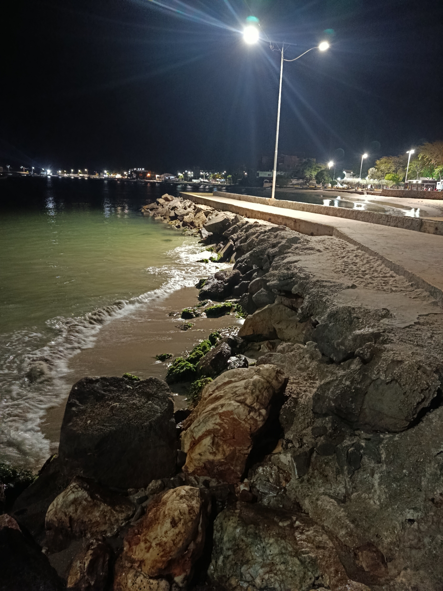 The image shows a nighttime scene in Juan Griego, Venezuela, featuring a stone breakwater extending into a body of water, likely the bay. The breakwater is constructed from large rocks and concrete. Waves are lapping against the rocky shoreline. A concrete walkway runs along the breakwater. Streetlights illuminate the scene. In the distance, lights from the town are visible along the waterfront. No people are visible in the image. Moss or algae grows on some of the rocks.