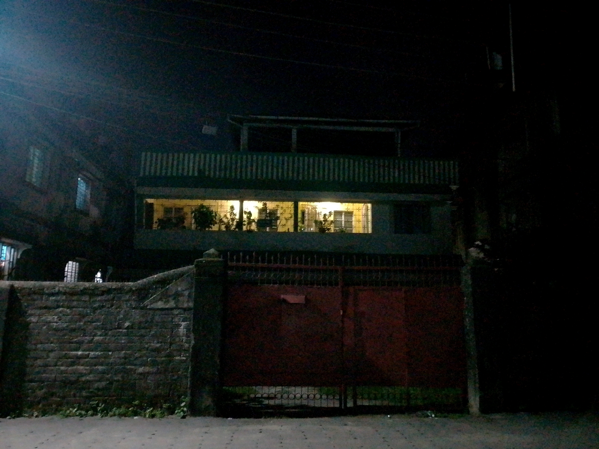 An exterior, nighttime, low-light, eye-level shot captures a dark brick wall and a large, dark red metal gate in Siliguri, India. The gate is ornate with a decorative metal grille along the top and appears to be partially open. To the left of the gate, the brick wall continues and is topped with what appears to be a concrete coping. Behind the wall and gate, a two-story building is visible. The ground floor has large windows, behind which plants and furniture can be seen, illuminated by interior lighting. The second floor features a balcony with vertical railings. The sky above is dark and indistinct, with faint hints of power lines. The scene is dark and atmospheric, suggesting a quiet, residential street at night.