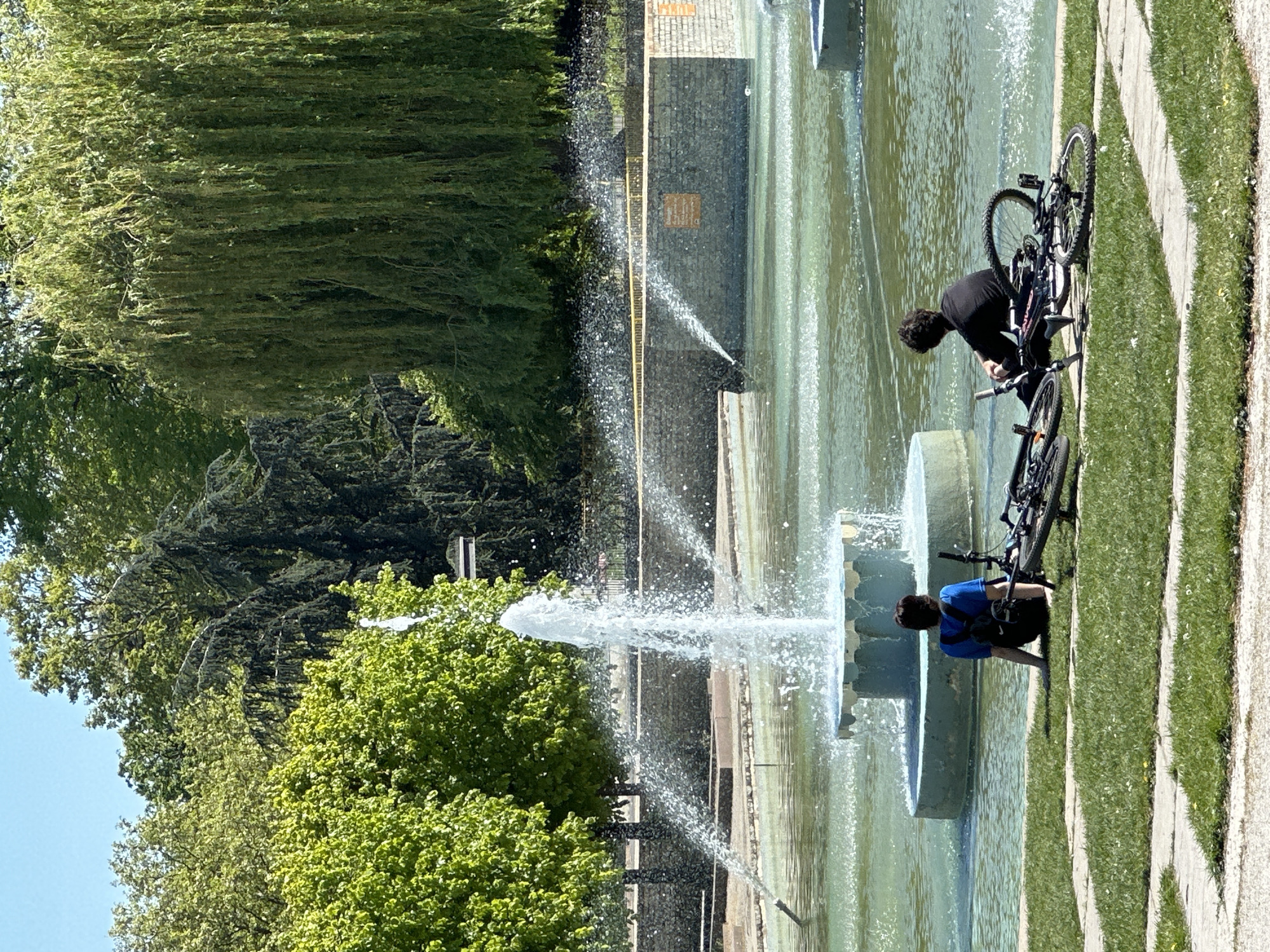 The photograph shows a fountain in a park. Two people are near the fountain with bicycles. One person, dressed in black, is standing near the fountain and appears to be interacting with one of the bicycles. The second person, dressed in blue, is sitting or crouching near the fountain. The fountain is made of multiple tiered circular structures, with water shooting from the top and sides. Trees and vegetation are visible in the background. The location is London, United Kingdom.