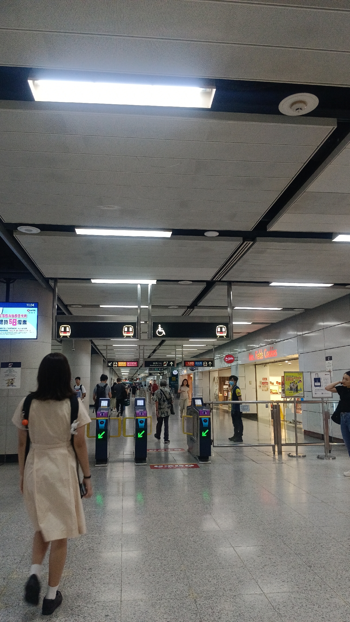 The image depicts a scene inside a MTR station in Hong Kong. A young woman wearing a light-colored dress and carrying a backpack is walking away from the camera, towards a set of ticket gates. Several other people are also visible, some walking through the gates and others standing. The environment is a typical MTR station interior, with tiled floors, fluorescent lighting, and signage indicating directions and services. There are also advertisements visible on screens and in shop windows. The ticket gates are automated, with green arrows indicating open pathways. The overall atmosphere suggests a busy public transportation hub during daylight hours.