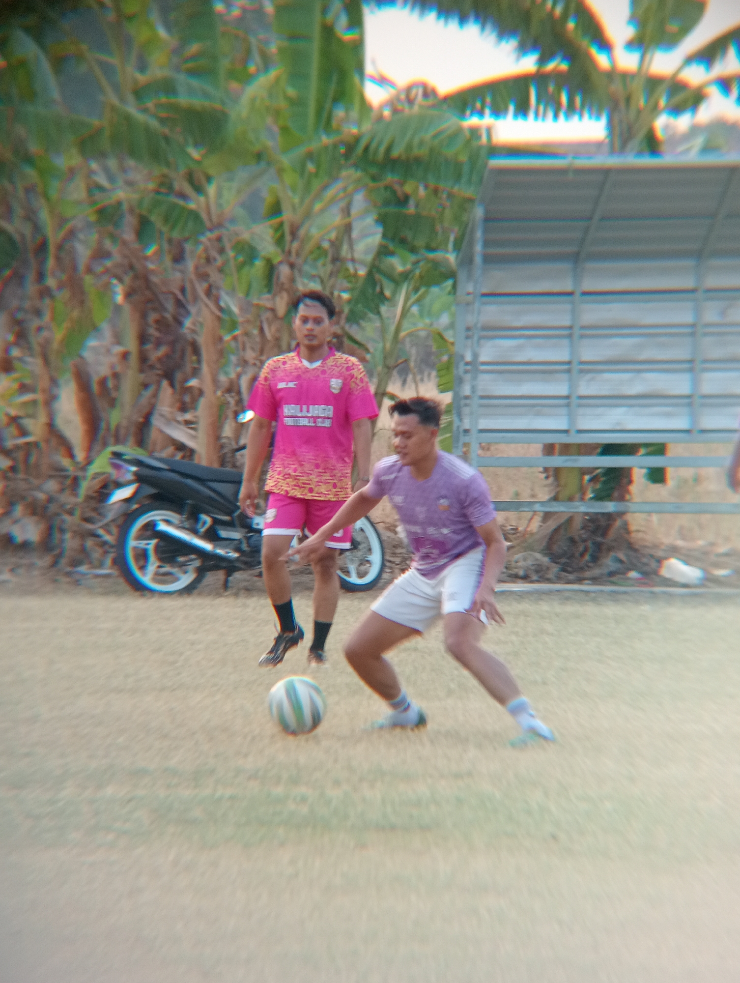 Two young men are playing soccer on a dirt field in Kenep, Indonesia. The man in the foreground, wearing a purple and white jersey and white shorts, is actively dribbling the ball. The man in the background, wearing a pink jersey and pink shorts, stands watching. A motorcycle is parked behind him. The environment appears to be a rural or semi-rural area, with palm trees and a simple metal structure visible. The image has a slightly blurred and vibrant quality, possibly due to motion or image processing.