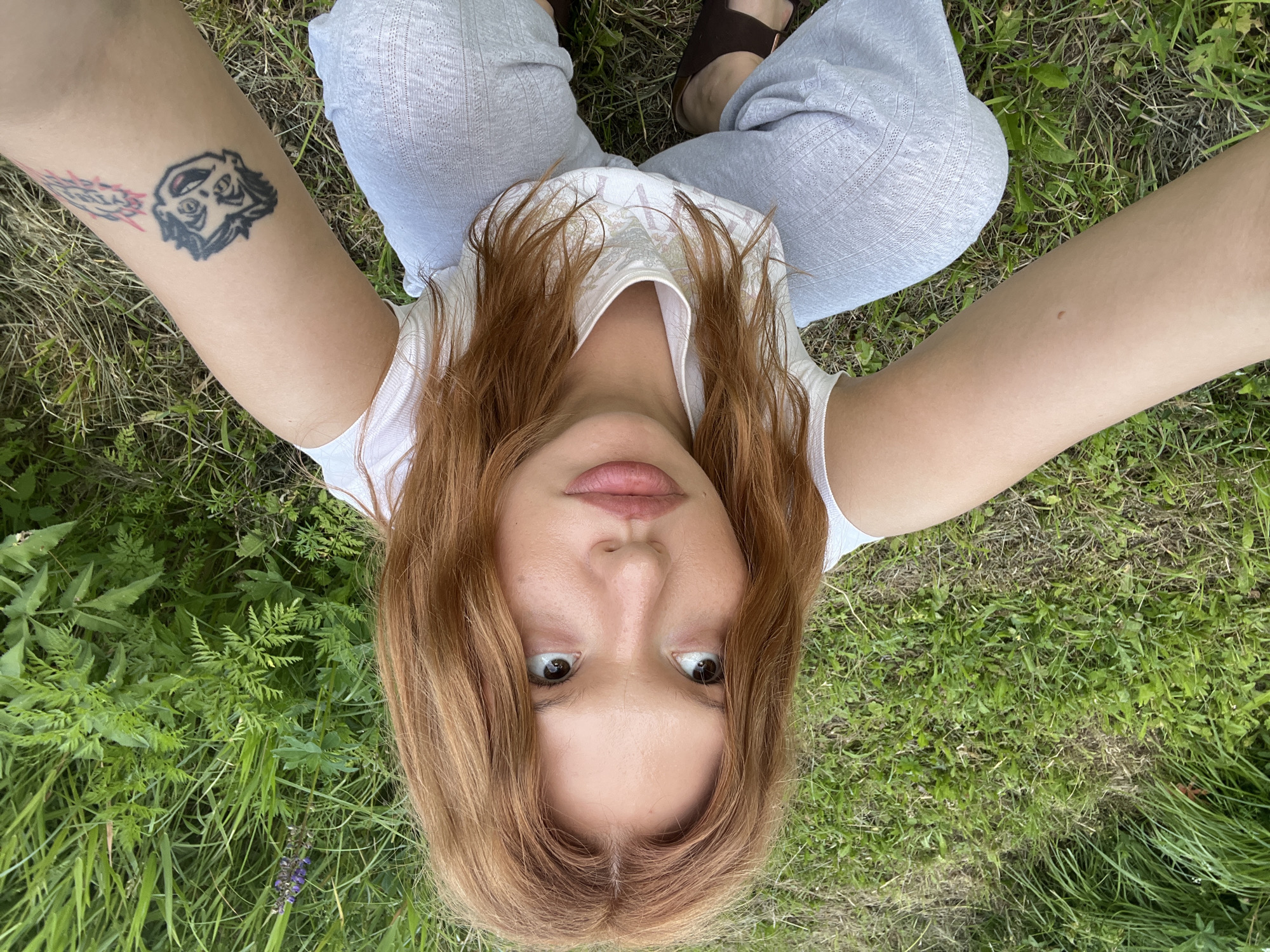 A young woman with long, reddish-brown hair and fair skin is crouching on a grassy area, looking up and to her left. She is wearing a white tank top and light gray patterned pants. A tattoo of a cartoonish face is visible on her right arm. The background is filled with lush green grass and foliage, suggesting an outdoor, possibly rural or park-like setting. There are no identifiable landmarks or structures that would indicate a specific location in Суроватиха, Russia. The lighting suggests it is daytime, likely under natural sunlight.