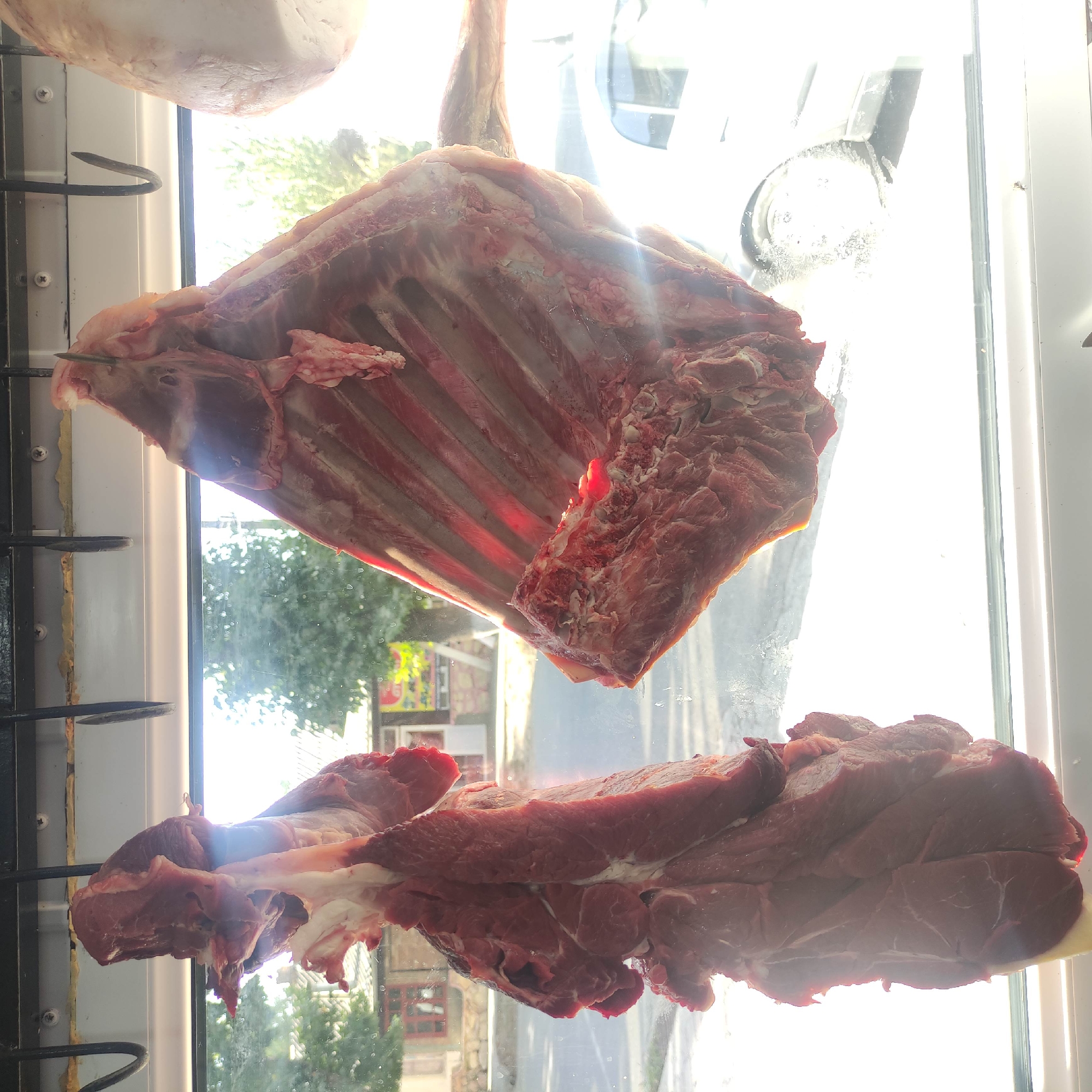 In Razzakov, Kyrgyzstan, two large cuts of raw meat hang from hooks inside what appears to be a display window, possibly at a butcher shop or market. The meat is dark red and appears to be on the bone in at least one of the cuts. The scene is brightly lit, suggesting it is daytime, with sunlight streaming in and creating lens flare and reflections on the glass. Outside the window, trees and buildings are visible, indicating an urban or suburban setting. A street with a car can also be seen through the glass, further confirming the location is outdoors. The overall impression is of a commercial establishment displaying fresh meat for sale.