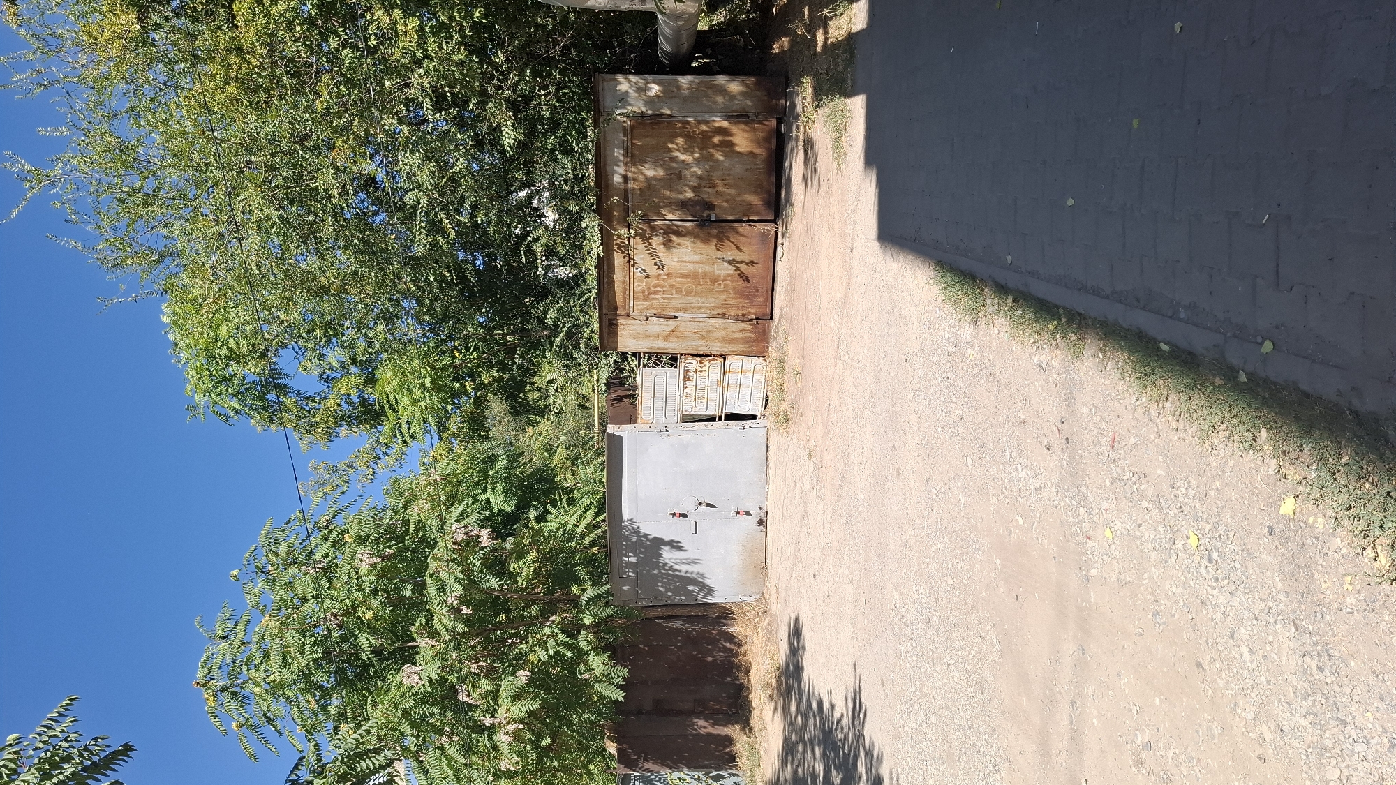 The image depicts a row of metal garages under a clear blue sky in Shymkent, Kazakhstan. The garages appear to be old and weathered, with varying degrees of rust and wear. One garage is a light grey color with red accents on its handles, while the others are predominantly brown and rusty. Between the grey garage and one of the rusty garages, there are several stacked units that resemble old radiators or air conditioning units.
The garages are situated on a gravel and dirt lot, with a paved walkway or sidewalk running along the lower right edge of the frame. Green trees and foliage are visible behind the garages, providing shade and a natural backdrop. The overall impression is one of a somewhat aged and utilitarian area, possibly a residential or industrial garage complex. There are no people visible in the image.