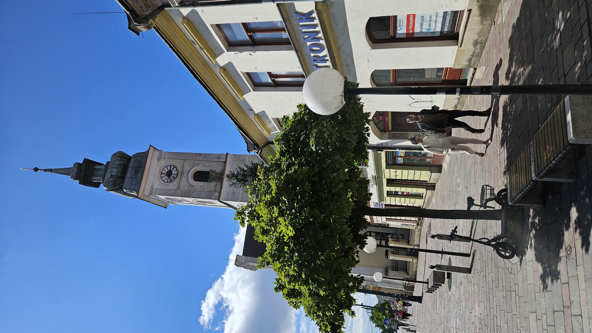 The image captures a street scene in Prešov, Slovakia, with a prominent clock tower as the central architectural element. A white building with yellow trim and the word "TRONIK" in blue lettering is on the right. Two individuals, a woman and a man, are walking hand-in-hand on the cobblestone sidewalk in front of the building. The woman is wearing a white sweater and light-colored pants, while the man is in a dark jacket and pants, with a baseball cap.
In the foreground, a parked electric scooter is visible next to a tree. Benches are placed along the sidewalk, and lampposts with globe lights are also present. The sky is blue with scattered white clouds. The clock tower, a tall structure with a spire, is visible behind the tree and other buildings. Shops with various signs are lined up along the street. The scene appears to be set during the daytime, with sunlight casting shadows on the ground.