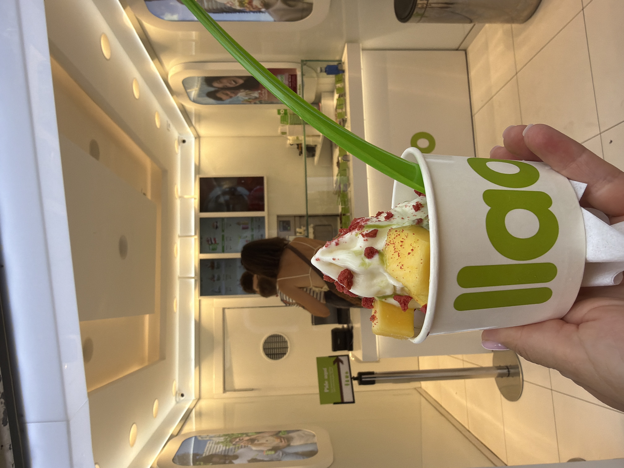 In an indoor establishment, likely a frozen yogurt shop, a hand is holding a white paper cup filled with frozen yogurt. The cup has green lettering that reads "llaollao". The frozen yogurt is topped with pieces of what appear to be mango and dried red berries. A bright green plastic spoon is inserted into the yogurt. In the background, the interior of the shop is visible, with a counter, menu displays on the wall, and several customers and employees. The lighting appears to be artificial and bright, with recessed ceiling lights and strip lighting along the counter. The floor is tiled.