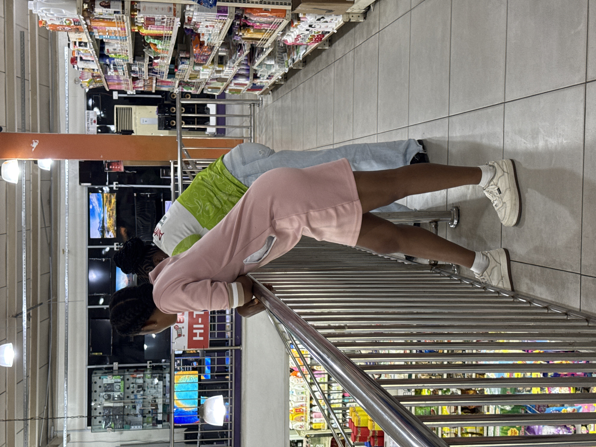 Two individuals are observed in what appears to be a retail environment in Ado, Nigeria. The foreground subject, dressed in a pink top, is leaning over a chrome railing, seemingly looking at items on shelves below. The second individual, wearing a green and white top and blue jeans, stands behind them, also looking down. The shelves are stocked with a variety of goods. In the background, televisions are displayed on a wall, and various electronic items are showcased. The flooring is tiled, and overhead lighting illuminates the scene.