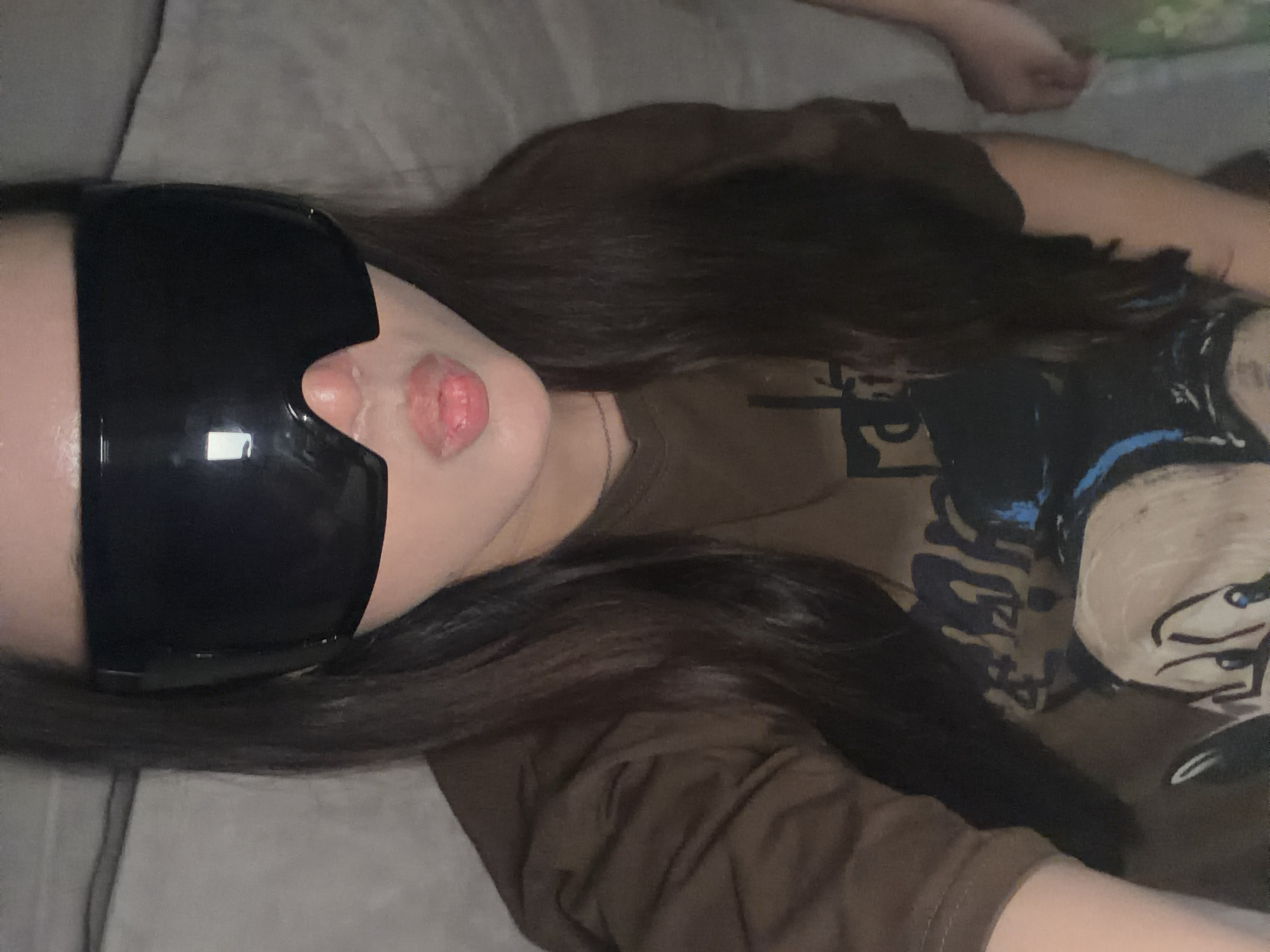 A woman is lying on a gray surface, likely a bed or couch, as indicated by the folds and textures of the material. She is wearing large, black, opaque sunglasses that cover her eyes and a portion of her face. Her lips are slightly puckered. The woman has long, dark brown hair that is spread out around her head. She is wearing a brown t-shirt with a graphic print on it. The location is Unknown, Unknown.
