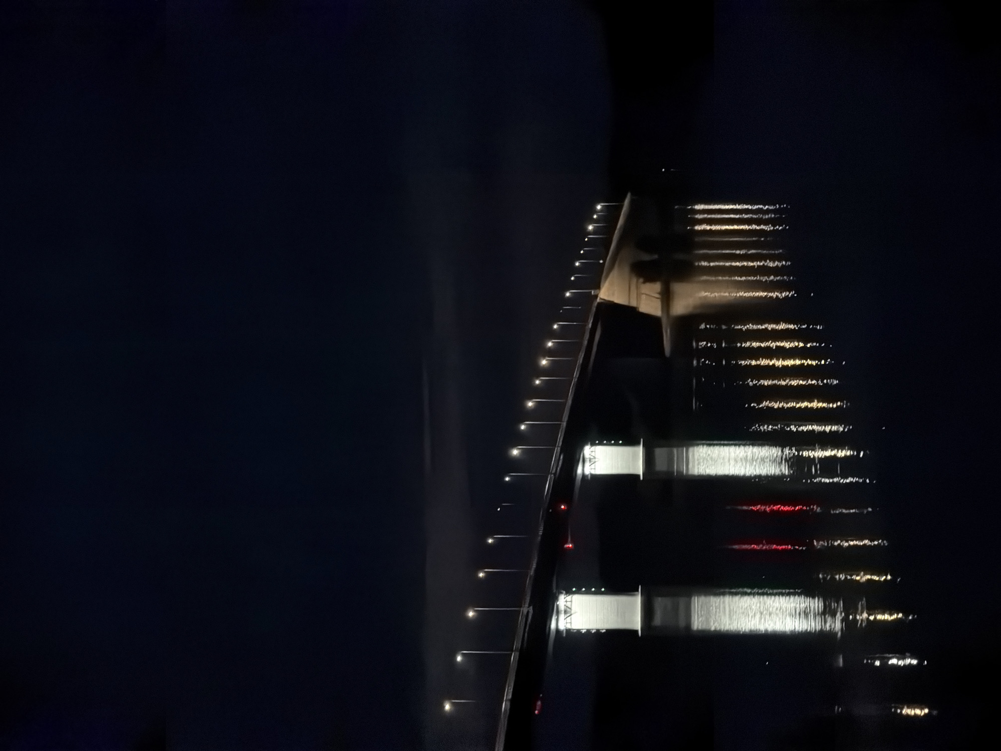 The image depicts a bridge illuminated at night, spanning across a body of water. The bridge has a long, elevated structure with rows of streetlights casting a warm glow along its length. Two prominent, brightly lit white support structures are visible beneath the bridge. The dark water below reflects the lights of the bridge, creating a shimmering effect. The background is a deep, dark sky, suggesting nighttime in a rural or less urbanized area. There are no people or vehicles visible in the image. The scene is serene and captures the architectural presence of the bridge against the night sky. This bridge is located in Tarasovo, Russia.