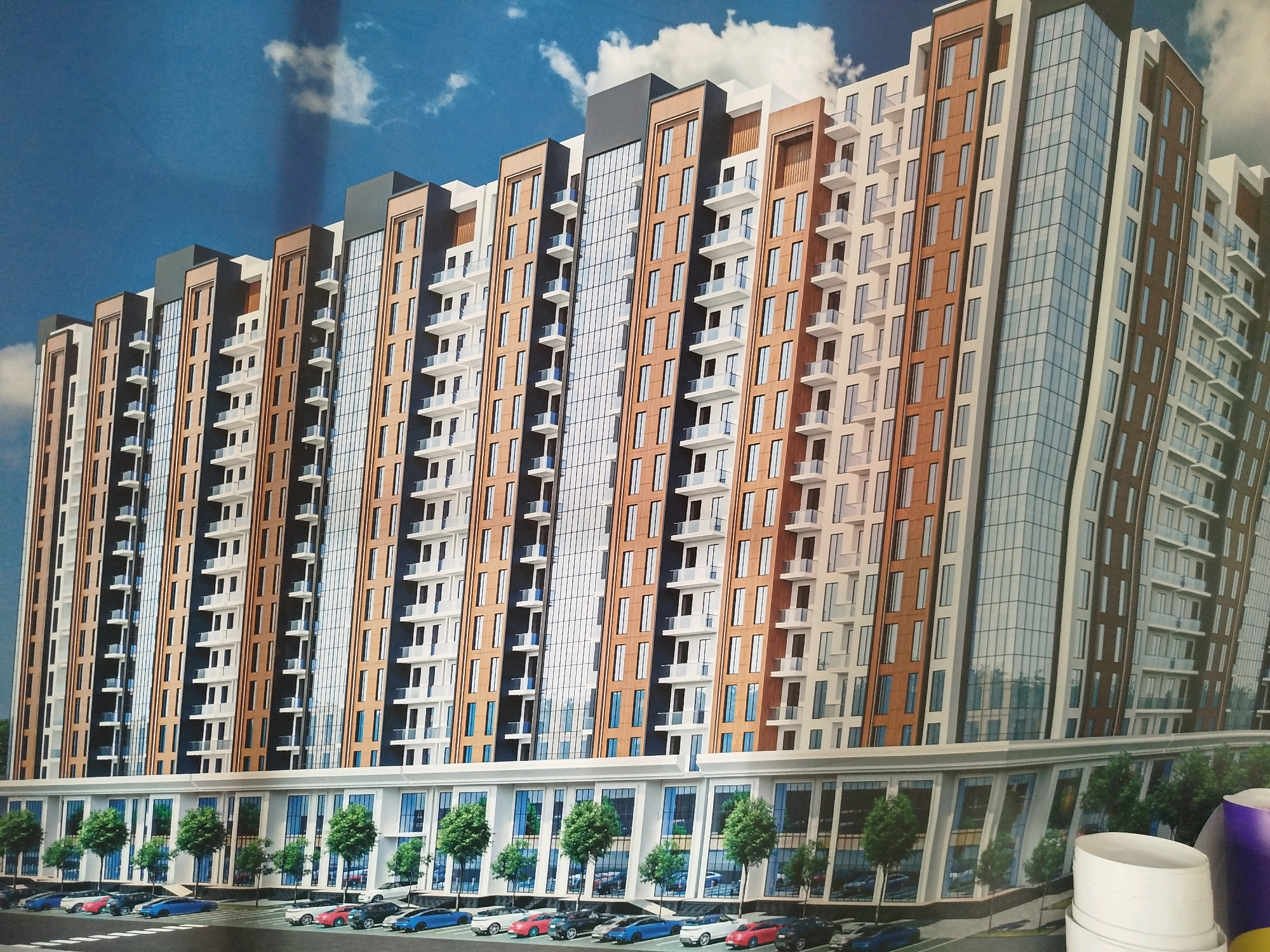 The image displays a rendering of a modern high-rise residential building with a white and brown facade, interspersed with large glass windows and balconies. The building features a tiered design with setbacks on upper levels and a prominent glass-covered central section. The lower level of the building is designed with a commercial or retail space featuring large windows and decorative columns. In front of this lower level, a landscaped area with trees and a parking lot filled with various cars, including sedans and SUVs, is depicted. The sky above is blue with scattered white clouds. The location is identified as Tashkent, Uzbekistan, and the building appears to be a new development.