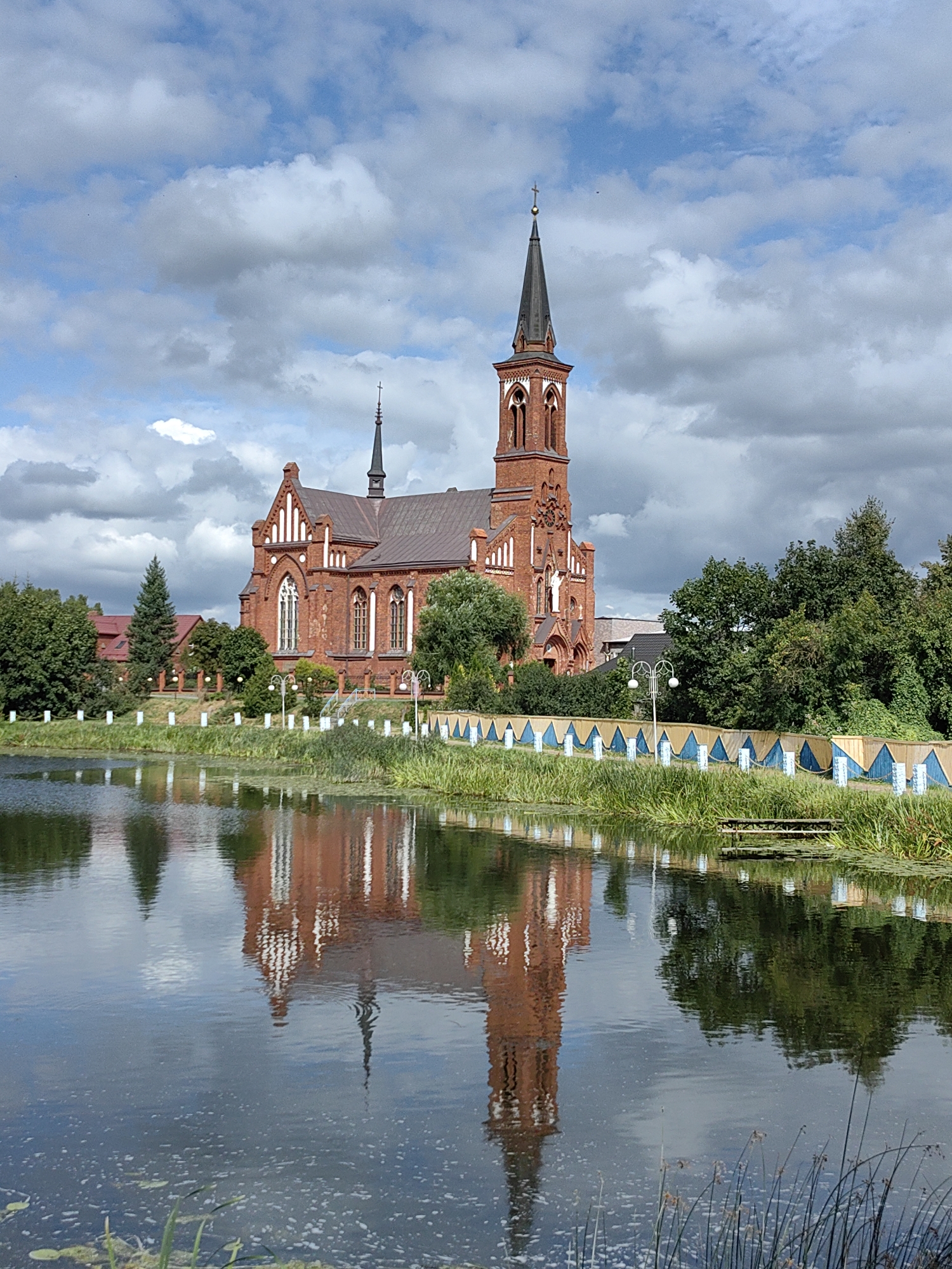 A red brick Neo-Gothic church with a tall, pointed steeple is reflected in a calm body of water in front of it. The church has arched windows and a dark brown roof. Green trees and a cloudy blue sky are also visible in the background. The body of water, likely a lake or river, is bordered by lush green grass and reeds. A wooden bench is situated on the edge of the water, and a decorative fence runs along the embankment. The scene appears to be from the city of Pastavy, Belarus, judging by the architectural style and typical landscape of the region.