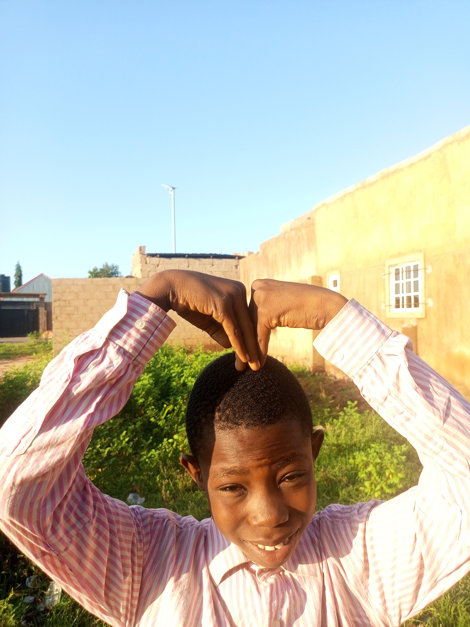 In Tempure, Nigeria, a young boy with dark skin and short, dark hair is photographed from the chest up. He is wearing a light pink and white striped button-down shirt. His left arm is bent upwards, with his hand resting on the top of his head, fingers curled. His right arm is also bent upwards, with his hand touching his left hand, forming a heart shape above his head. He is smiling broadly, with his teeth visible. The background features a building with a light yellow facade and a white-paned window. In the distance, a solar-powered street light is visible against a clear blue sky. The ground is covered in green foliage and some scattered debris.