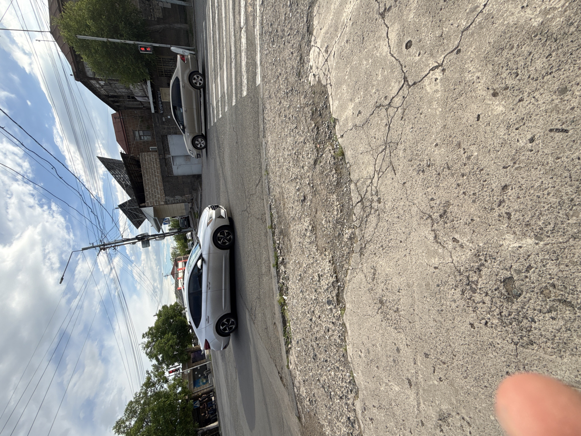 An outdoor, eye-level, daytime scene depicts an urban street environment with cars, buildings, and utility infrastructure under a partly cloudy sky.
In the immediate foreground, a blurry human finger is partially visible in the bottom-left. The ground consists of heavily cracked and worn light-grey concrete pavement, transitioning into a rougher, gravelly patch with sparse weeds.
The midground features a paved asphalt road. A silver Hyundai Elantra sedan is in motion, heading from left to right. Further right, a gold-colored Volkswagen Polo sedan is either stopped or moving slowly at a white zebra pedestrian crossing.
In the background, on the left, several green deciduous trees partially obscure buildings. One visible building has a dark facade and a large wall graphic or mural. Two indistinct individuals are seated or standing on a low wall or bench on the sidewalk. On the right, a prominent utility pole is heavily laden with numerous black wires crisscrossing the frame, supporting a streetlamp. To the pole's right, an old, two-story building with a dark, weathered facade and a reddish-brown roof stands. Adjacent to it on the left is a building constructed of dark stone or brick, topped with a red roof and multiple large, dark, angular structures on its upper exterior, possibly solar panels. A red traffic light, displaying the digit "6," is mounted on a pole near the Volkswagen Polo and the older building. A small tree is situated near the traffic light pole.
The sky is partly cloudy, showing patches of blue sky between white clouds. The overall impression is an aged urban setting.