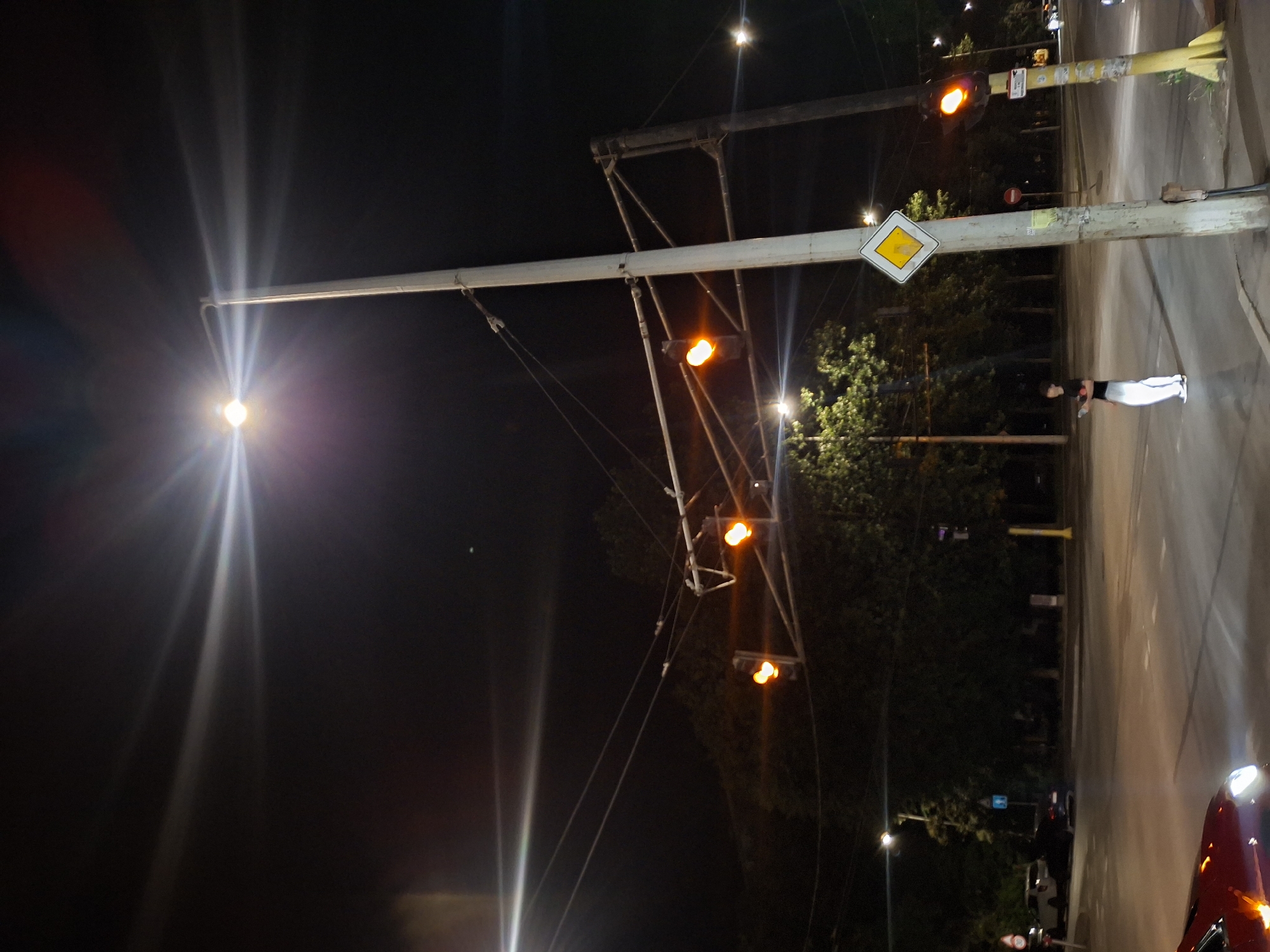 At night, a street intersection in Ruse, Bulgaria, is illuminated by a bright streetlamp and traffic signals. A young person stands on the sidewalk, holding a phone. Traffic lights hang above the intersection, displaying amber lights. A yellow sign with a diamond shape is mounted on a pole. In the foreground, a red car's headlights are visible. Trees line the background, and the road surface is wet, reflecting the lights.