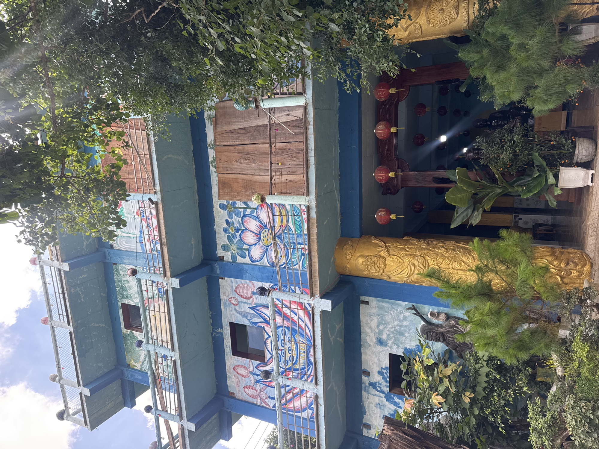 The image depicts the exterior of a multi-story building in Da Lat, Vietnam, characterized by its vibrant blue paint and ornate murals. The building features several balconies with railings adorned with decorative elements and strings of small lights. The walls are decorated with colorful paintings of lotus flowers and other floral motifs, as well as what appear to be mythical or spiritual figures. Large wooden double doors are visible on one of the upper levels.
At the ground level, the entrance is framed by large, golden, ornate pillars. Hanging above the entrance are rows of red, decorative lanterns. The area in front of the building is filled with lush greenery, including potted plants and trees. A statue of a figure resembling a wise elder or a shaman is partially visible among the foliage, positioned against the building's wall. The scene suggests a religious or cultural site, possibly a temple or a place of spiritual significance, set within the natural landscape of Da Lat. The sky above is partly cloudy, with sunlight filtering through the leaves of trees.