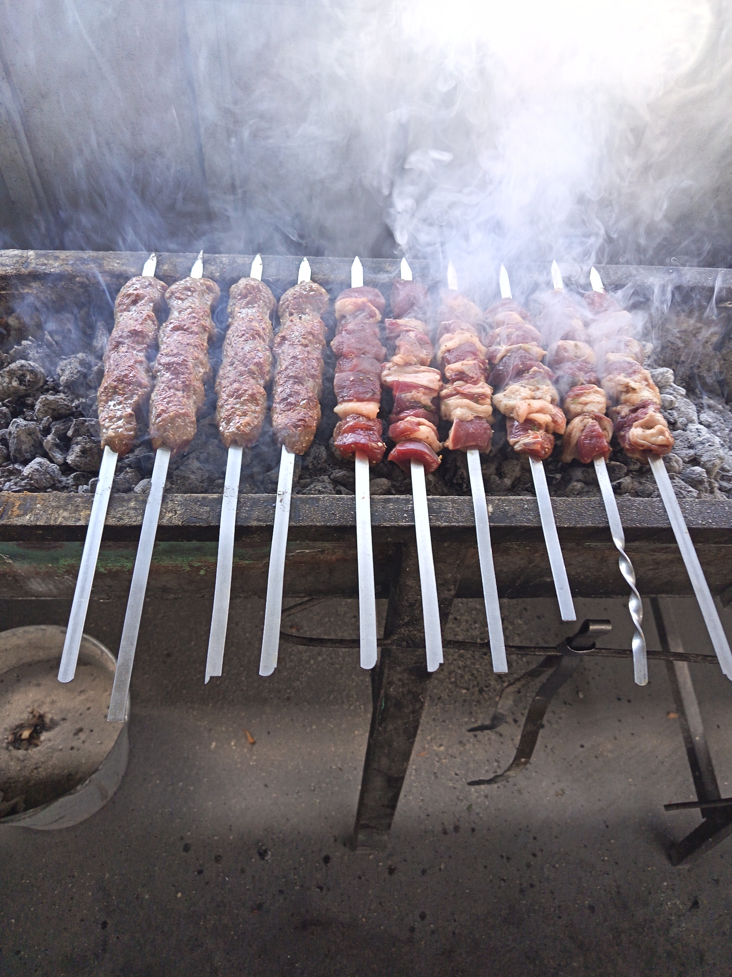 A close-up, high-angle shot shows multiple skewers of meat grilling over charcoal. The skewers are arranged in a row, with some appearing to be seasoned ground meat and others chunks of raw meat with fat. Smoke rises from the grill, obscuring the background. The grill itself appears to be made of metal and is set on a concrete surface. To the left, a metal bucket with ash and debris is visible. The overall scene suggests an outdoor cooking environment, possibly a street food stall or a backyard barbecue. The location is identified as Rizak, Uzbekistan.