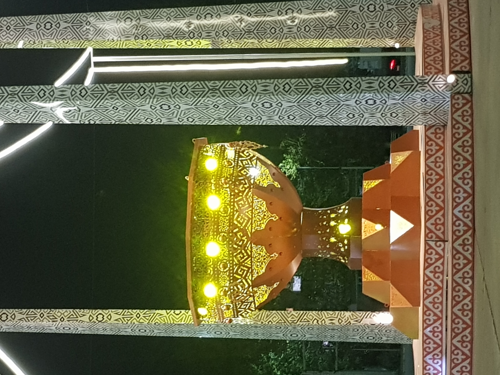 The image shows a large, ornate, golden brazier illuminated by yellow lights, situated in the city of Kurchatov, Kazakhstan. The brazier is supported by a multi-tiered, reddish-brown base with geometric patterns. The entire structure is framed by two tall, decorative pillars with intricate black and white geometric designs. White LED lights run vertically along the pillars and are also integrated into the structure of the brazier and its base. The background is dark, suggesting it is nighttime, with some greenery visible behind the main structure. The ground in the foreground has a patterned border. No people are visible in the frame.