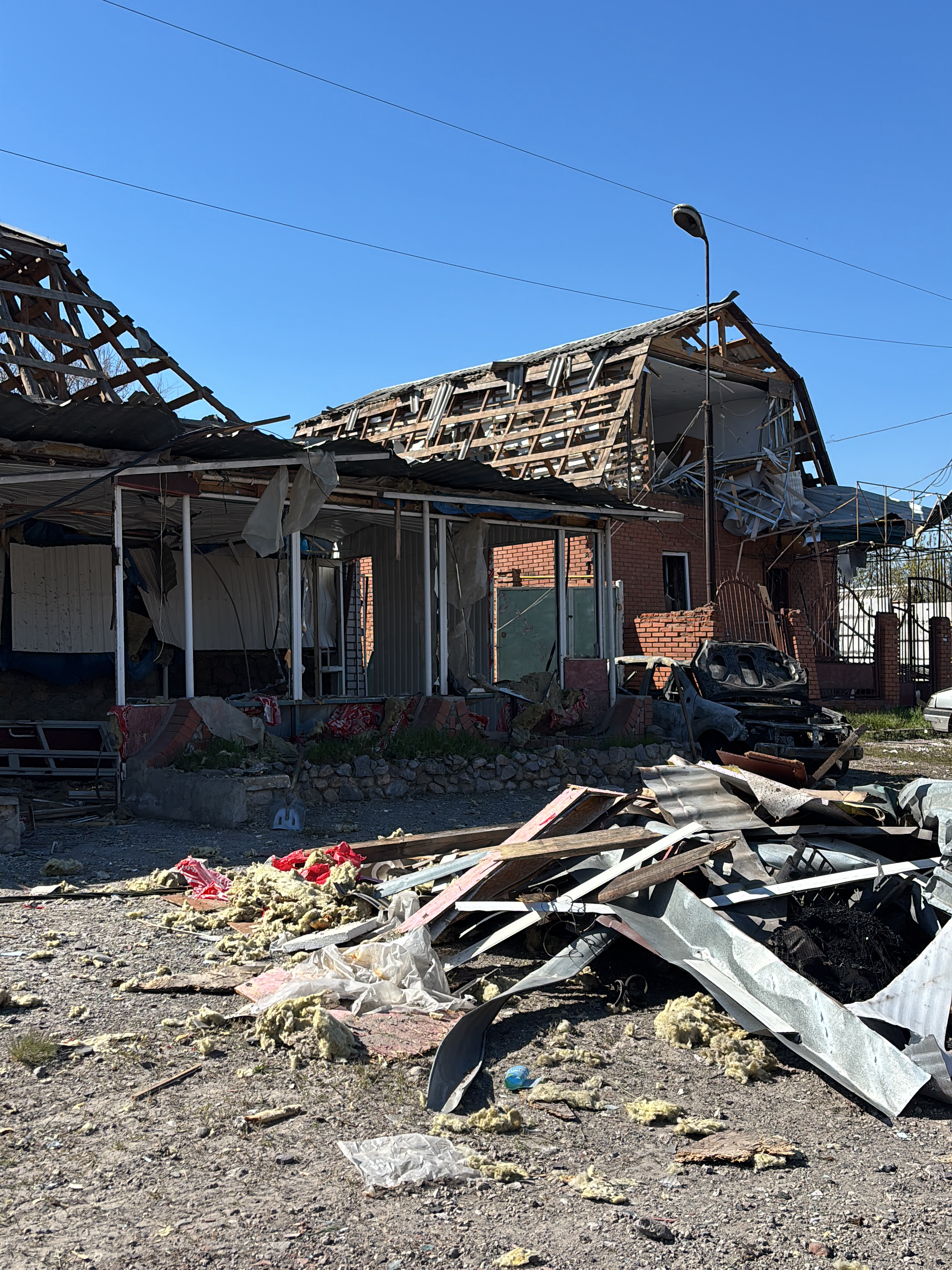The image shows a scene of devastation, likely the aftermath of a conflict, in Vysokyi, Ukraine. A partially destroyed brick building has a collapsed roof, exposing the wooden support structure. Nearby is what is left of another partially destroyed structure with a collapsing white portico and roof. Debris, including metal sheets, wooden planks, and insulation material, litters the ground. A burned-out car is visible next to the brick building. A street lamp stands behind the building, seemingly untouched, while powerlines run across the sky. The sky is clear and blue. There are no people visible in the image.