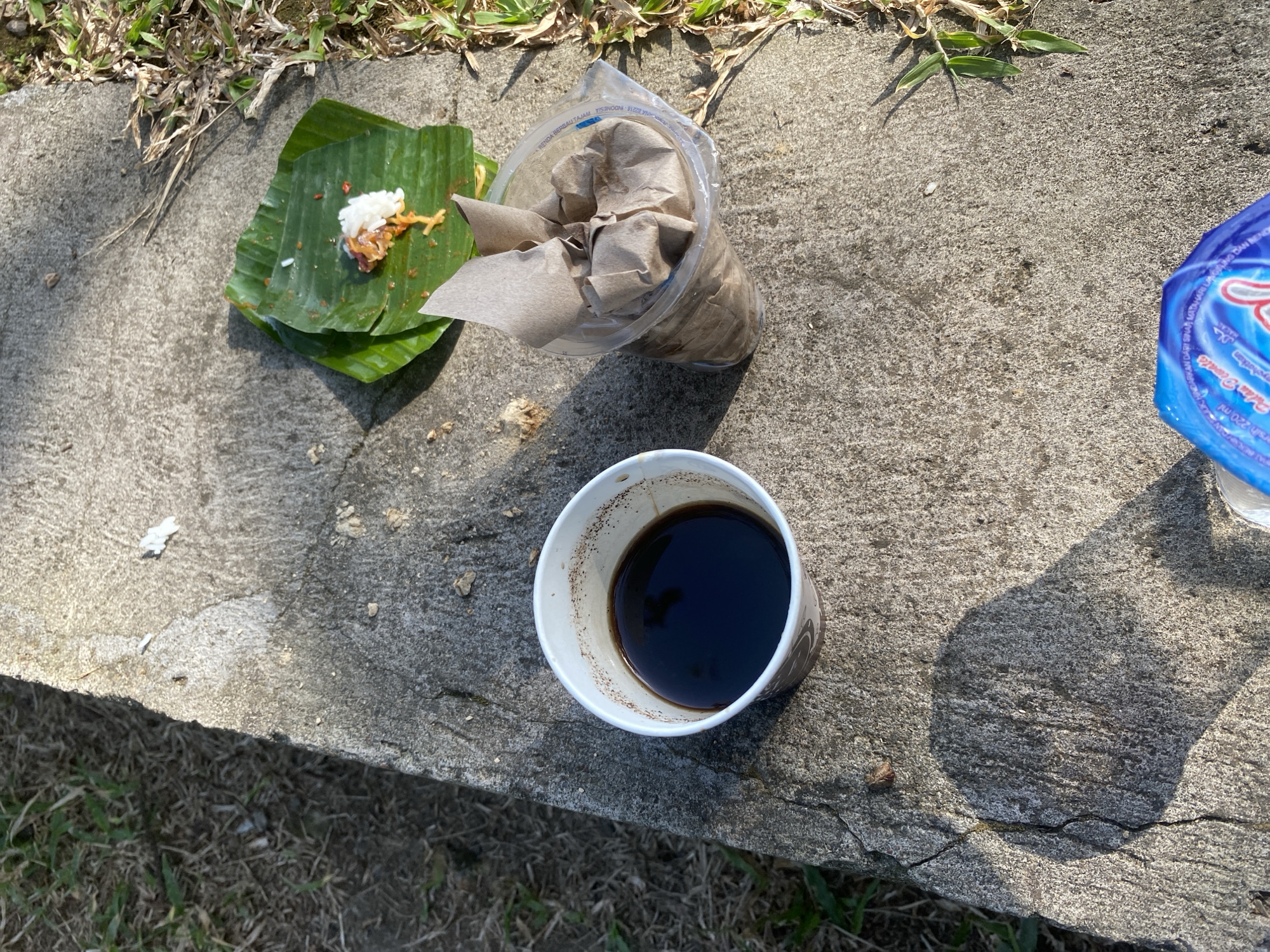 In Pekutatan, Indonesia, on a concrete surface adjacent to dry grass, a meal is presented outdoors. A white paper cup filled with dark liquid, likely coffee, sits to the left. To the right, a portion of food is served on green banana leaves, consisting of white rice and a small amount of what appears to be meat or chili-based topping. Adjacent to the food is a clear plastic cup containing a brown paper wrapper. The scene is illuminated by sunlight, casting shadows on the concrete. No individuals are present.