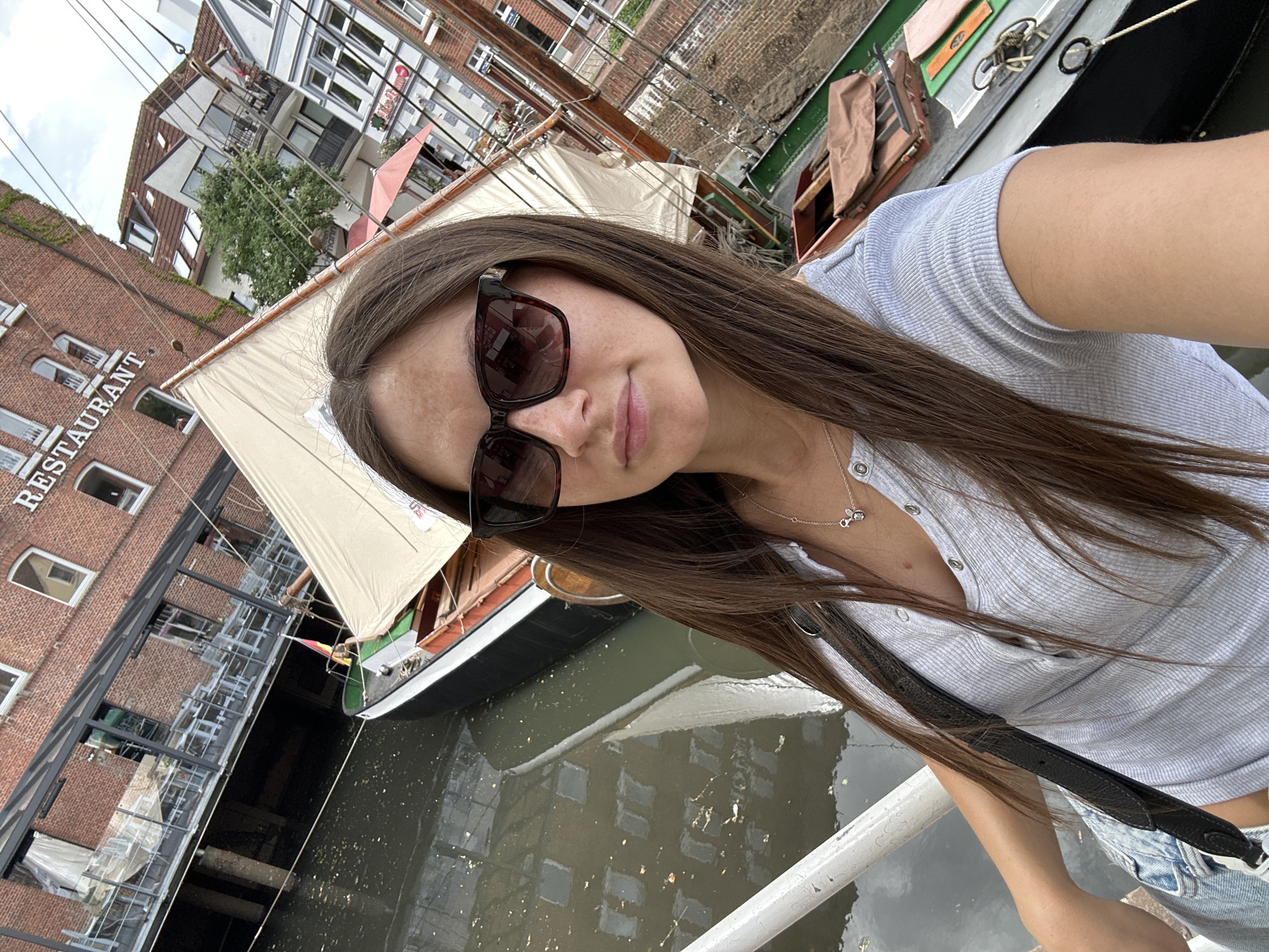 A woman is taking a selfie in Buxtehude, Germany. She is wearing sunglasses, a light blue shirt, and a necklace. A boat is docked next to her. Behind the boat is a building with "RESTAURANT" written on it. Water is visible between the woman and the building. Other buildings and boats are also in the background.