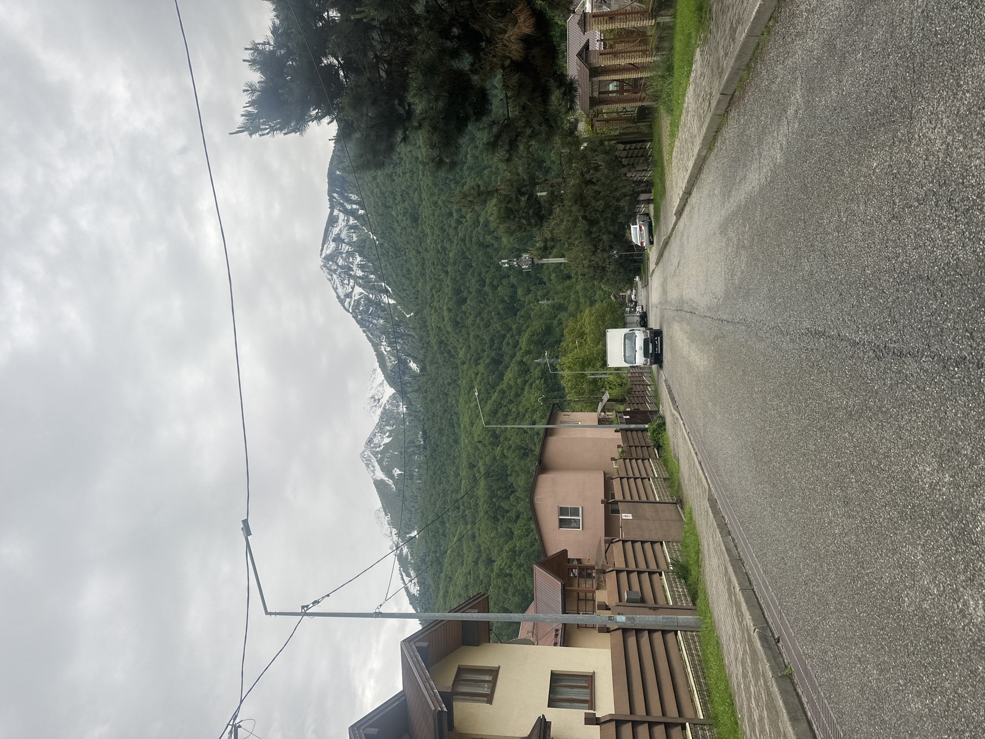 The image shows a street view in Krasnaya Polyana, Russia. The street is paved and lined with residential buildings on the right. A white van and other vehicles are visible on the street. A mountain covered in green trees with some snow-capped peaks is in the background. The sky is overcast. There are power lines running along the side of the road.