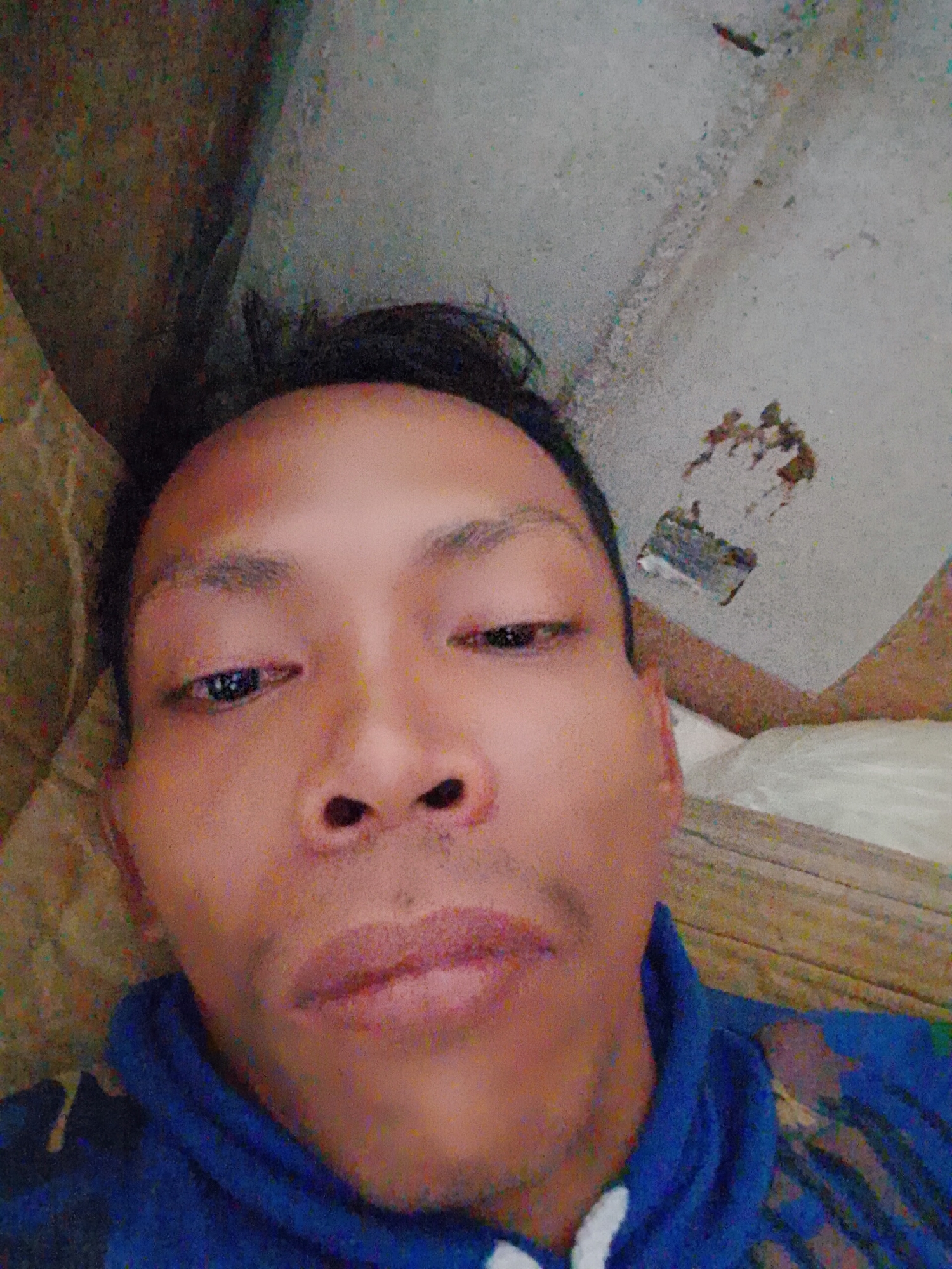 A close-up selfie of a person with brown skin, dark hair, and brown eyes, wearing a blue hoodie with a white neckline. The background includes a beige wall and a textured white ceiling with some damaged areas. The location is Bukit Ketok, Indonesia.