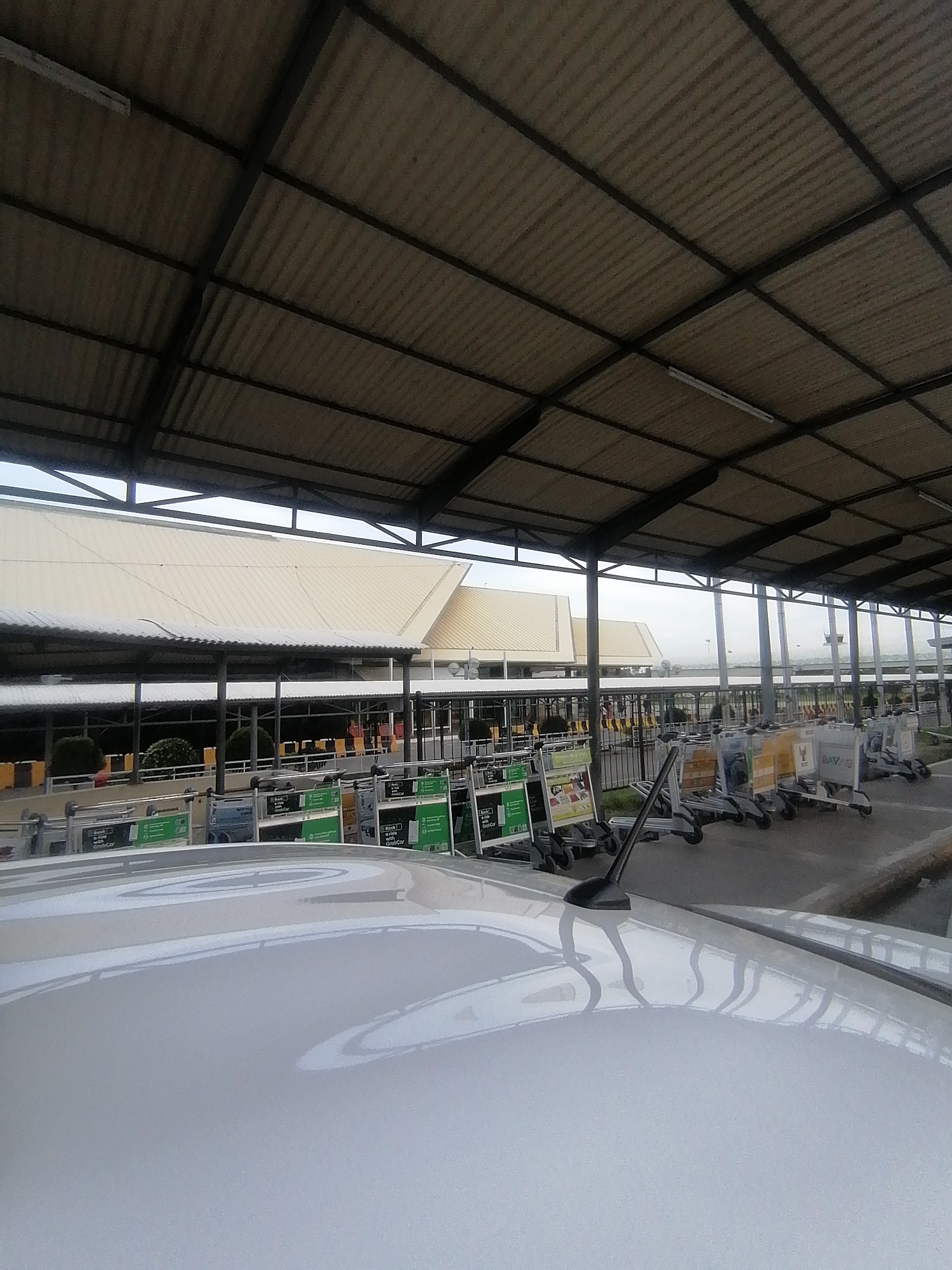 The image depicts a covered outdoor area, likely a drop-off or pick-up zone at an airport or a large transportation hub in Davao City, Philippines.
In the foreground, the shiny white roof of a car is prominently featured, with a black antenna visible on its right side. The car's polished surface reflects the overhead structure.
Above the car and extending across the upper portion of the image is a large, expansive canopy made of corrugated metal roofing supported by dark metal beams. Several fluorescent light fixtures are attached to the underside of this overhead structure.
Underneath the canopy, numerous luggage trolleys or baggage carts are lined up in rows. Many of these trolleys feature green promotional signs. Text visible on some of these signs clearly states "Book a ride with GrabCar," indicating a ride-hailing service is advertised or available. Other partial text like "BAV..." can be seen on some carts.
In the mid-ground, beyond the trolleys and under the shelter, a building with a light-colored, possibly off-white or beige, corrugated metal roof is visible, likely an airport terminal. Another similar building structure appears further in the background.
The scene is devoid of people, creating a quiet atmosphere. The lighting is diffused, suggesting an overcast sky or the deep shade provided by the large canopy overhead, consistent with daytime conditions. The overall impression is one of functionality and utility at a transportation facility.